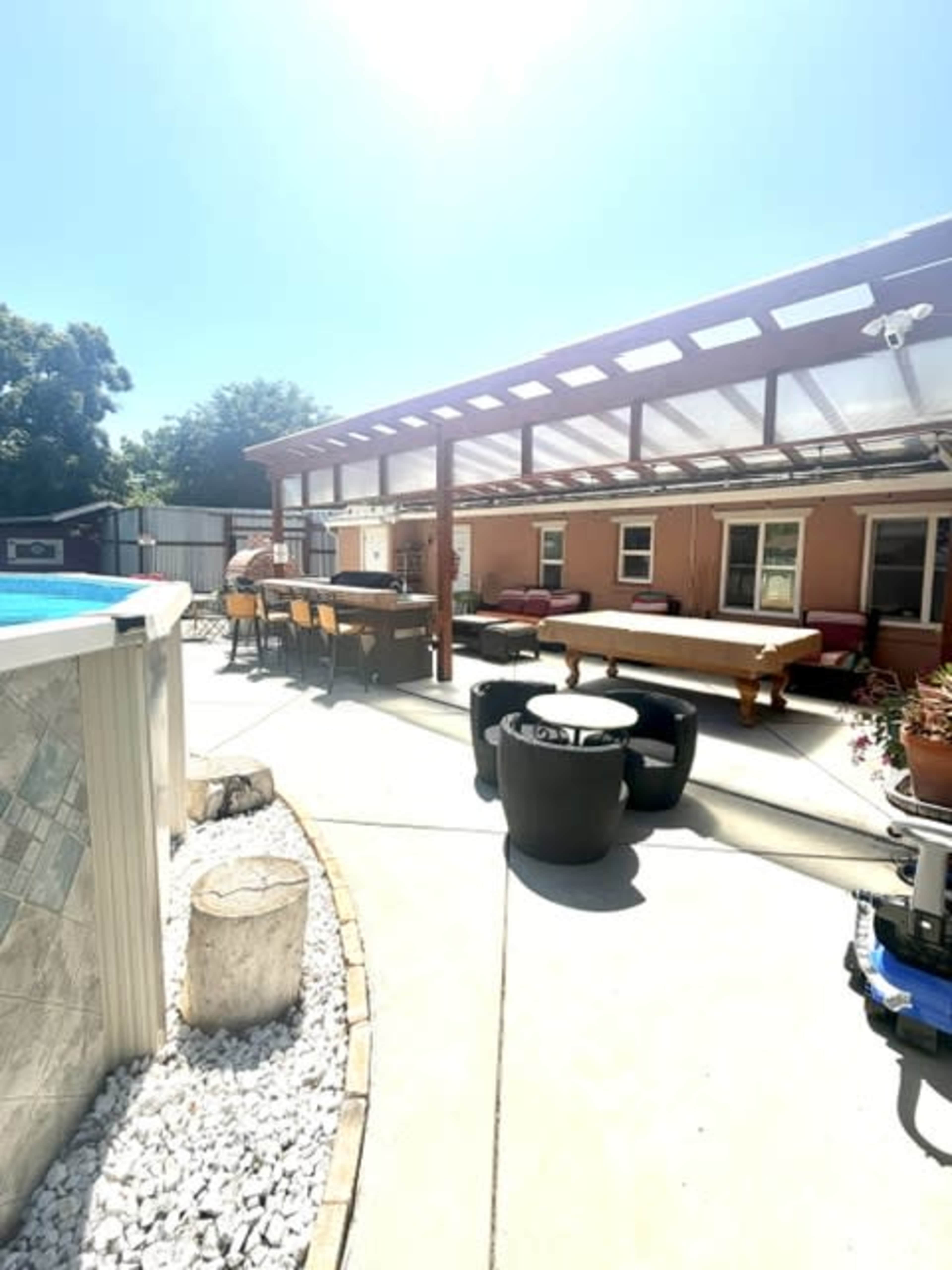 The image shows a well-lit patio area featuring a swimming pool, outdoor furniture, and a sheltered dining space.