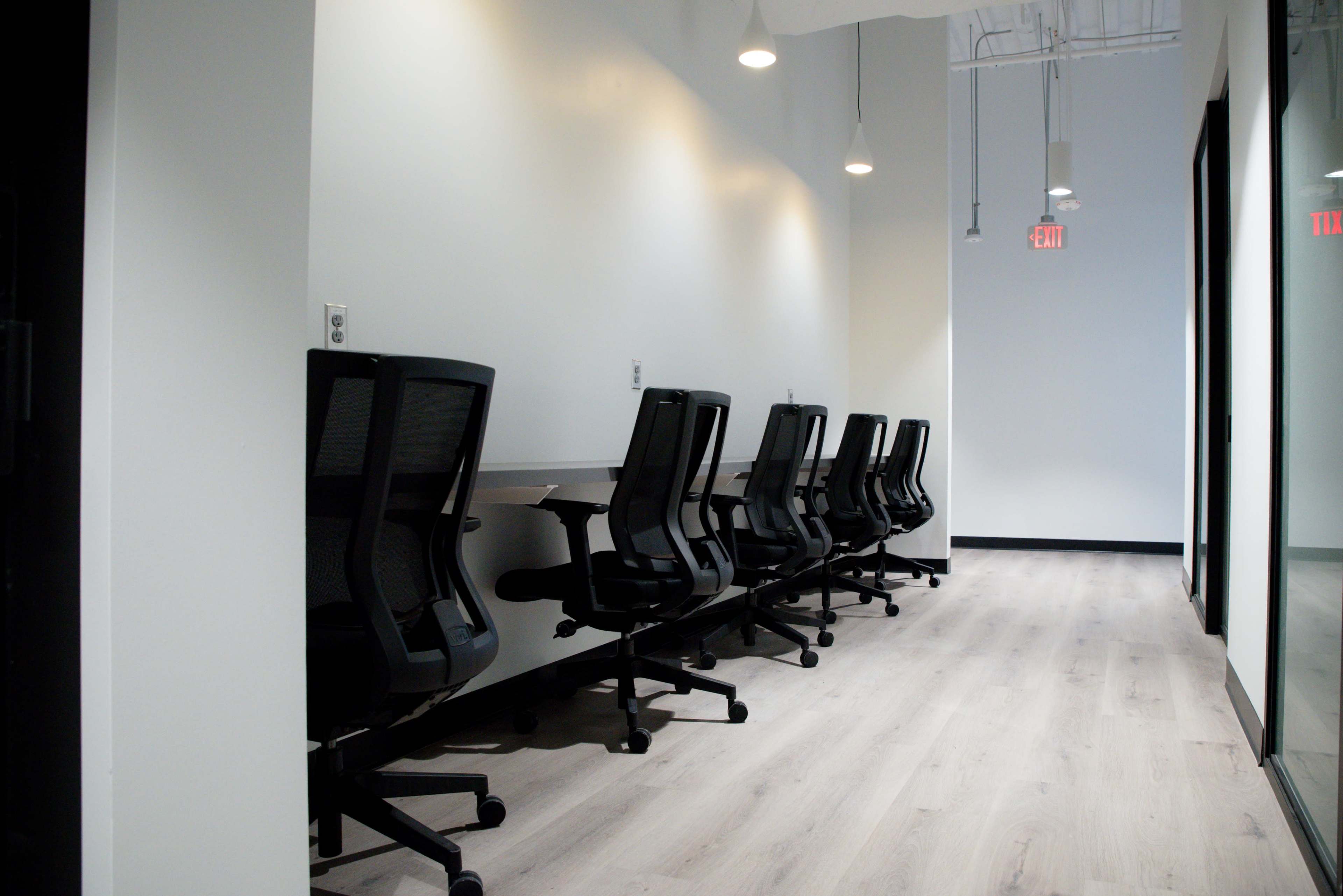 The image shows a row of black office chairs lined up at a long desk in a modern, minimalist hallway with white walls and hardwood flooring.