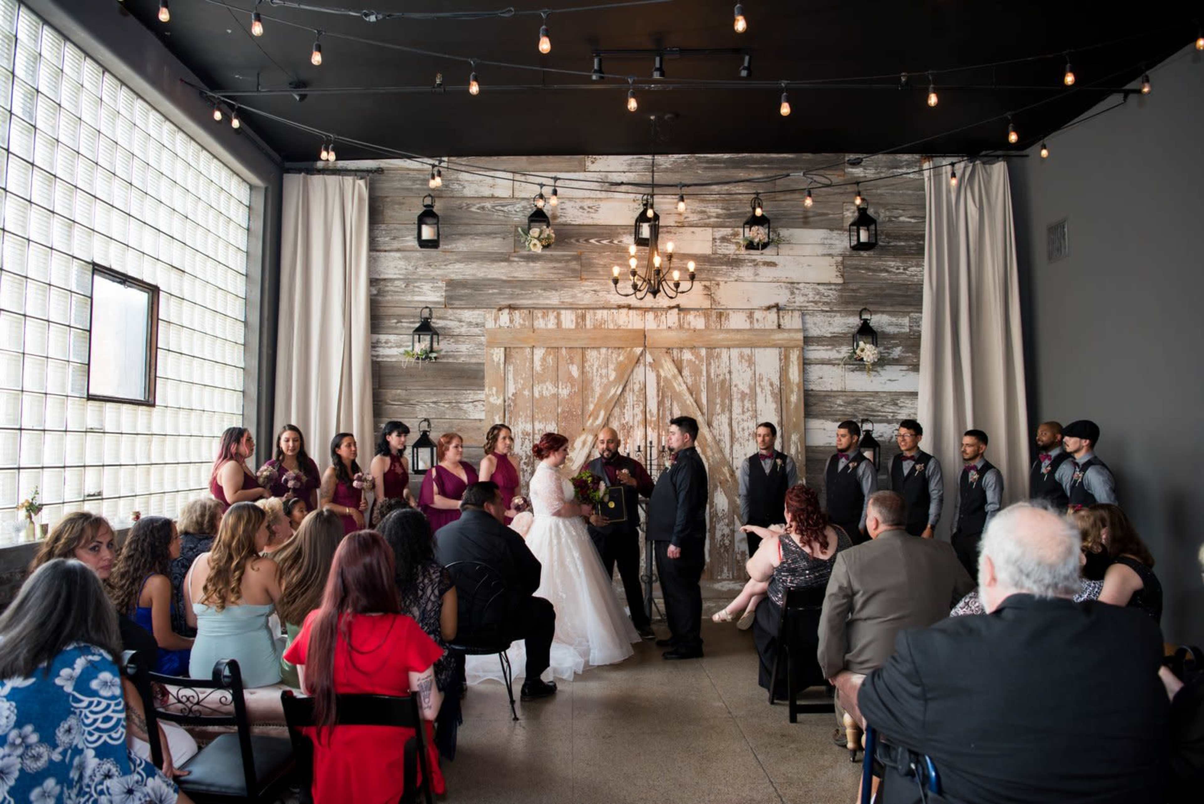 A wedding ceremony takes place indoors, with a couple standing at an altar and guests seated nearby, surrounded by rustic decor and string lights.