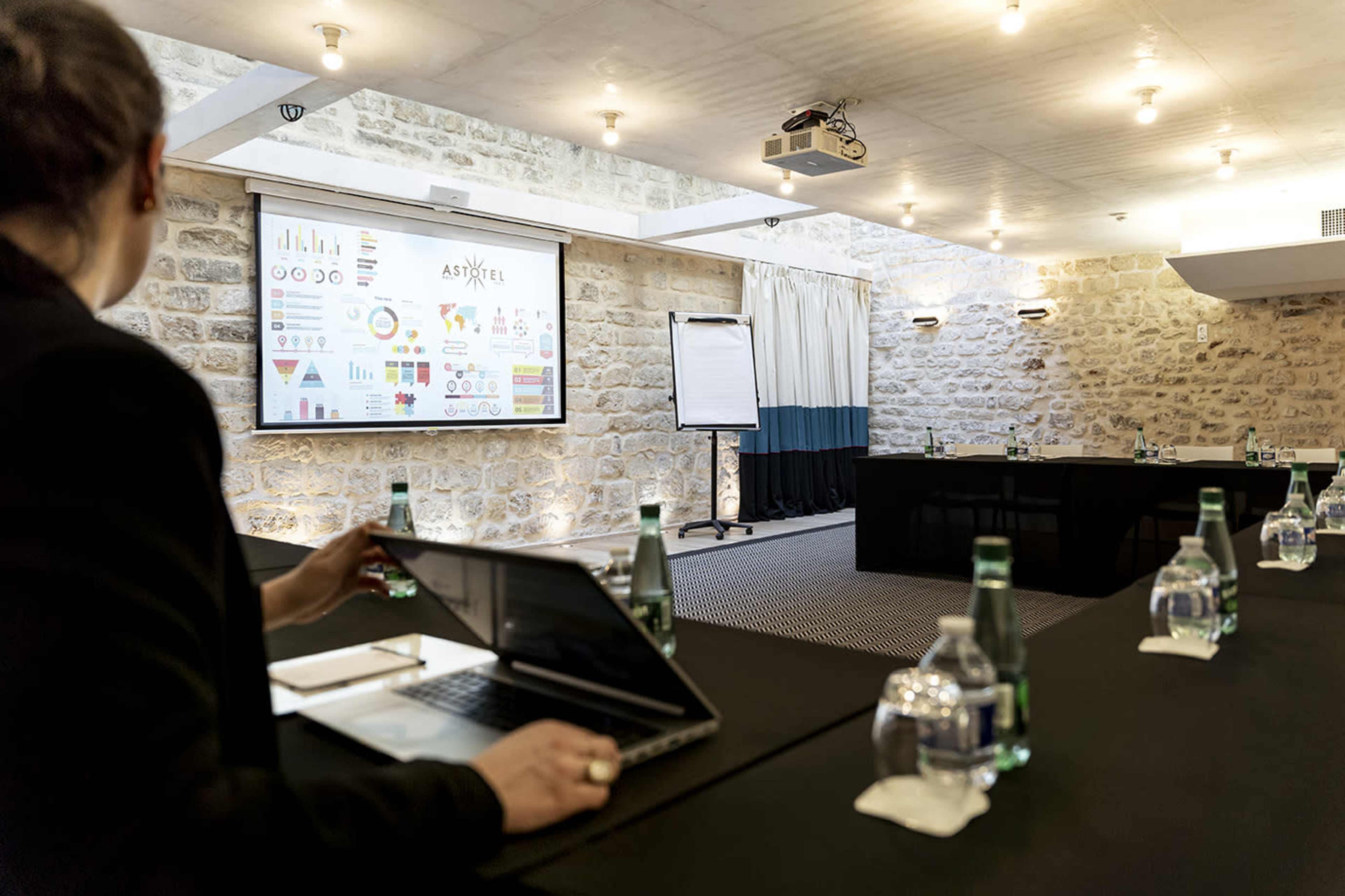 A person sits at a table in a conference room, viewing a presentation displayed on a screen, with multiple water bottles and laptops on the table.