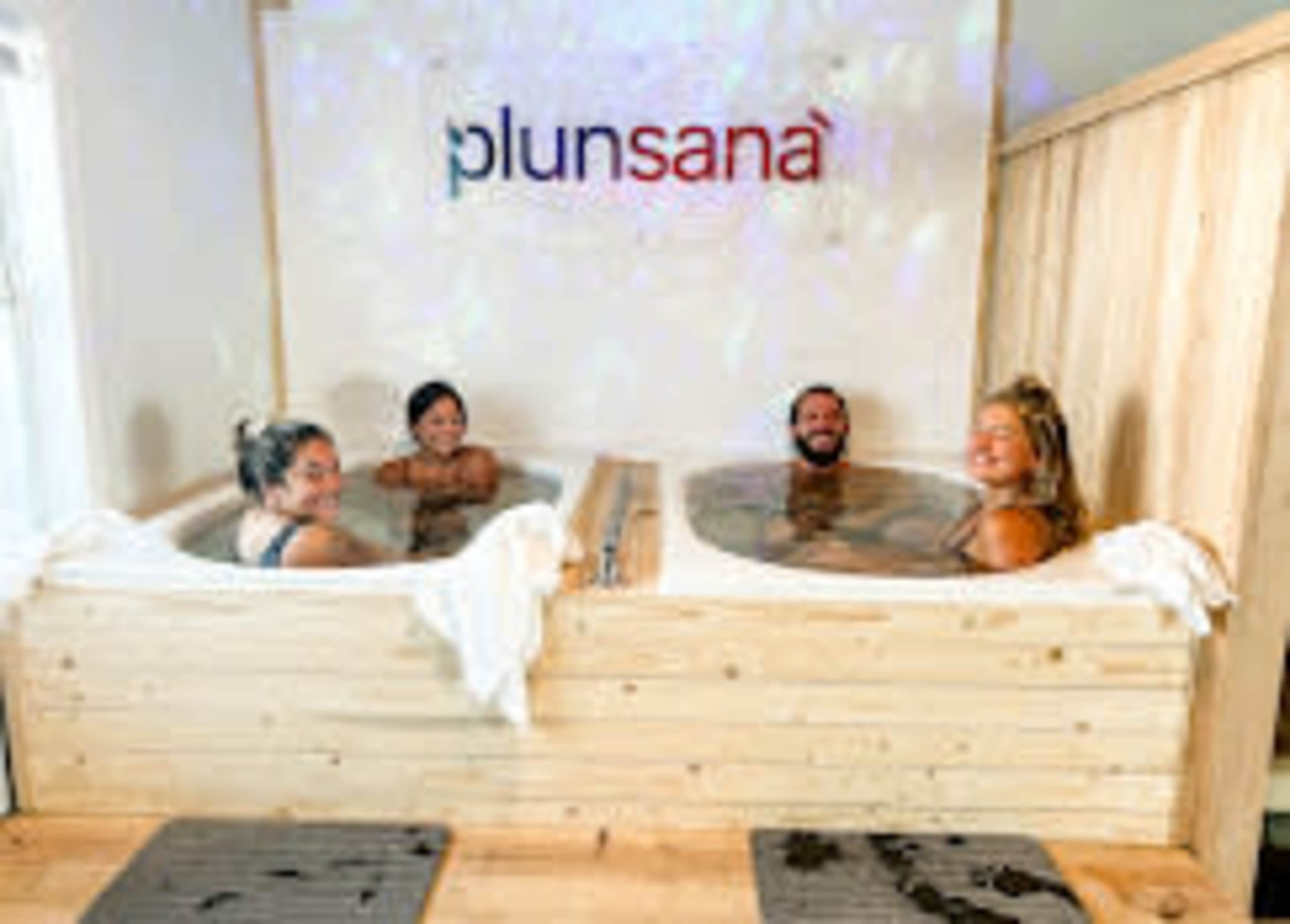 Four people are relaxing in wooden hot tubs at a wellness center with the sign "plunsana" in the background.