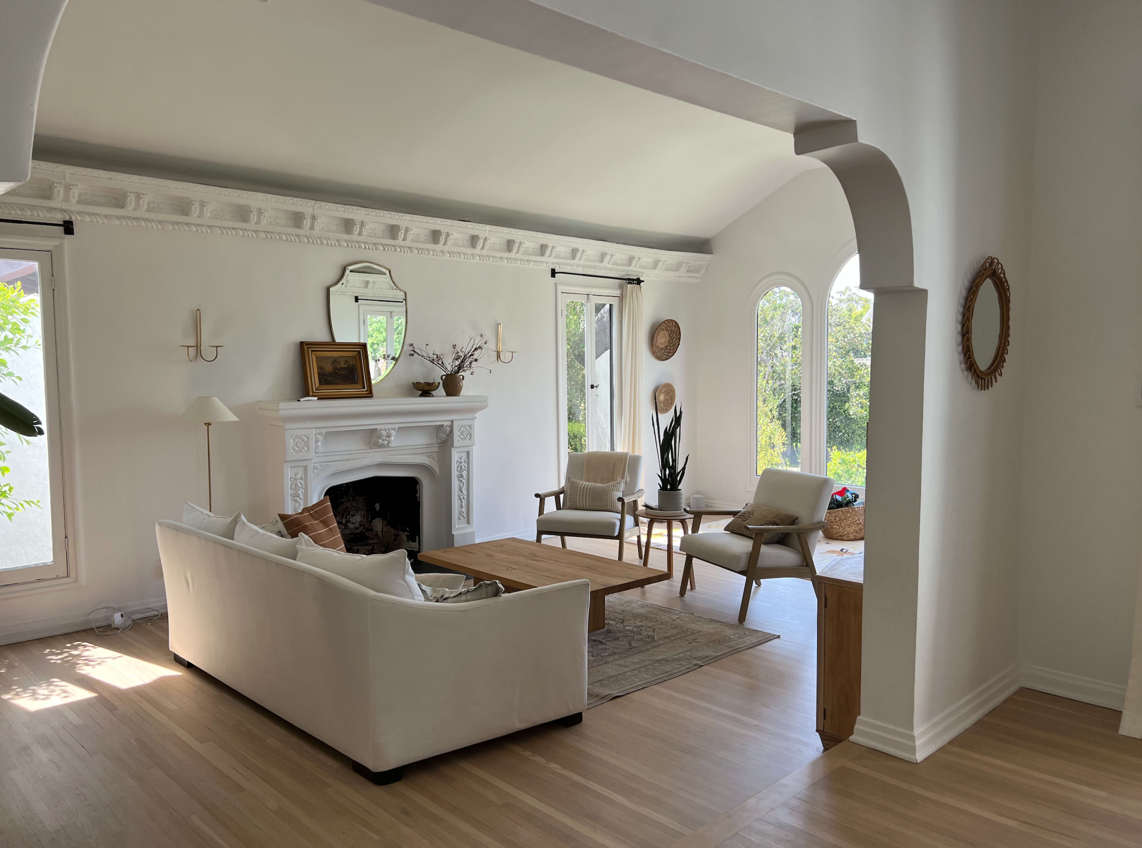 The image shows a bright living room with a white sofa, wooden coffee table, and large windows allowing natural light to fill the space.