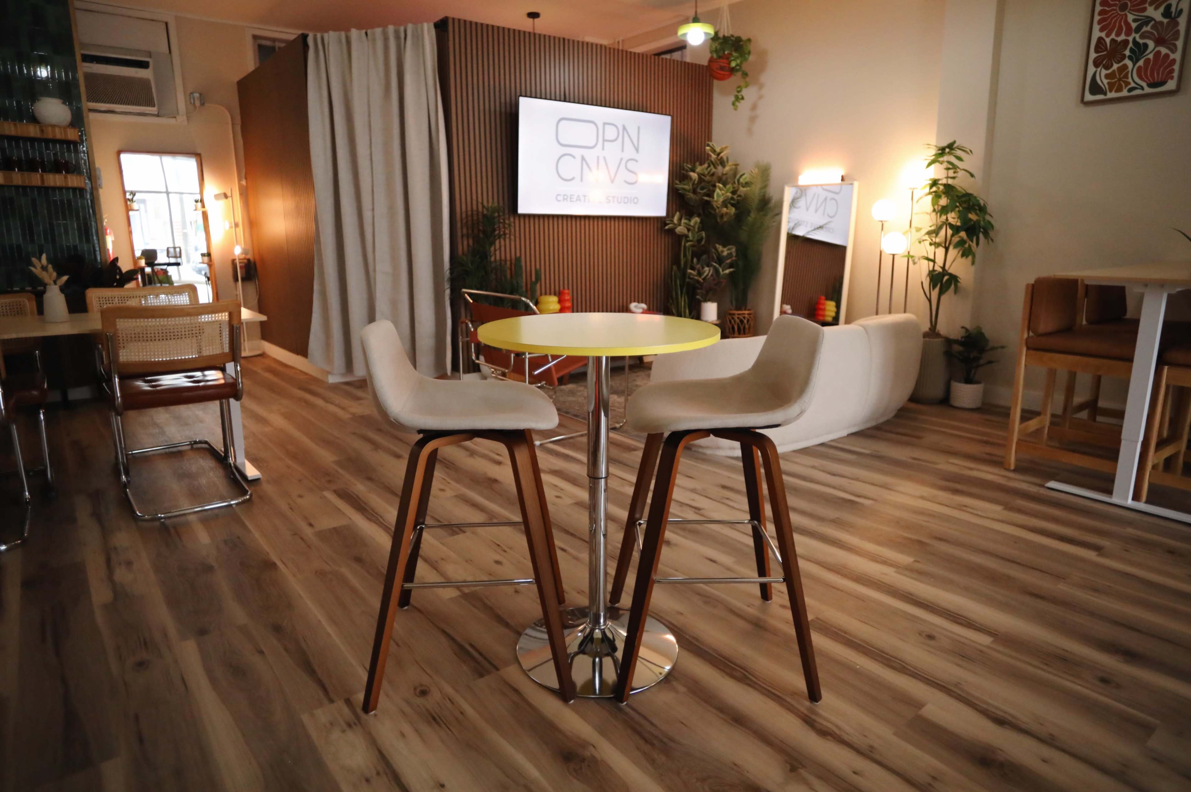 The image shows a modern interior of a workspace featuring a circular yellow table with two high stools, surrounded by wooden flooring and green plants.