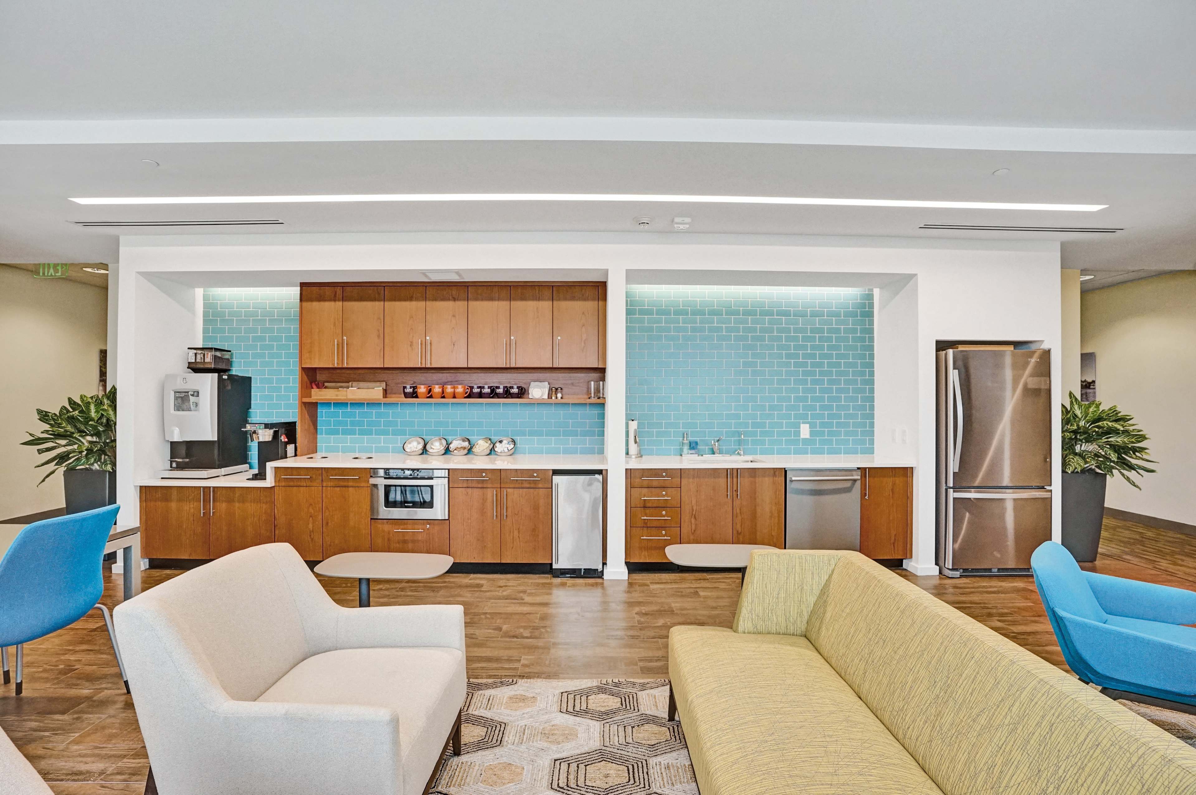 A modern kitchen area with wooden cabinets, a stainless steel refrigerator, a microwave, and seating arrangements featuring a gray sofa and a yellow patterned couch.