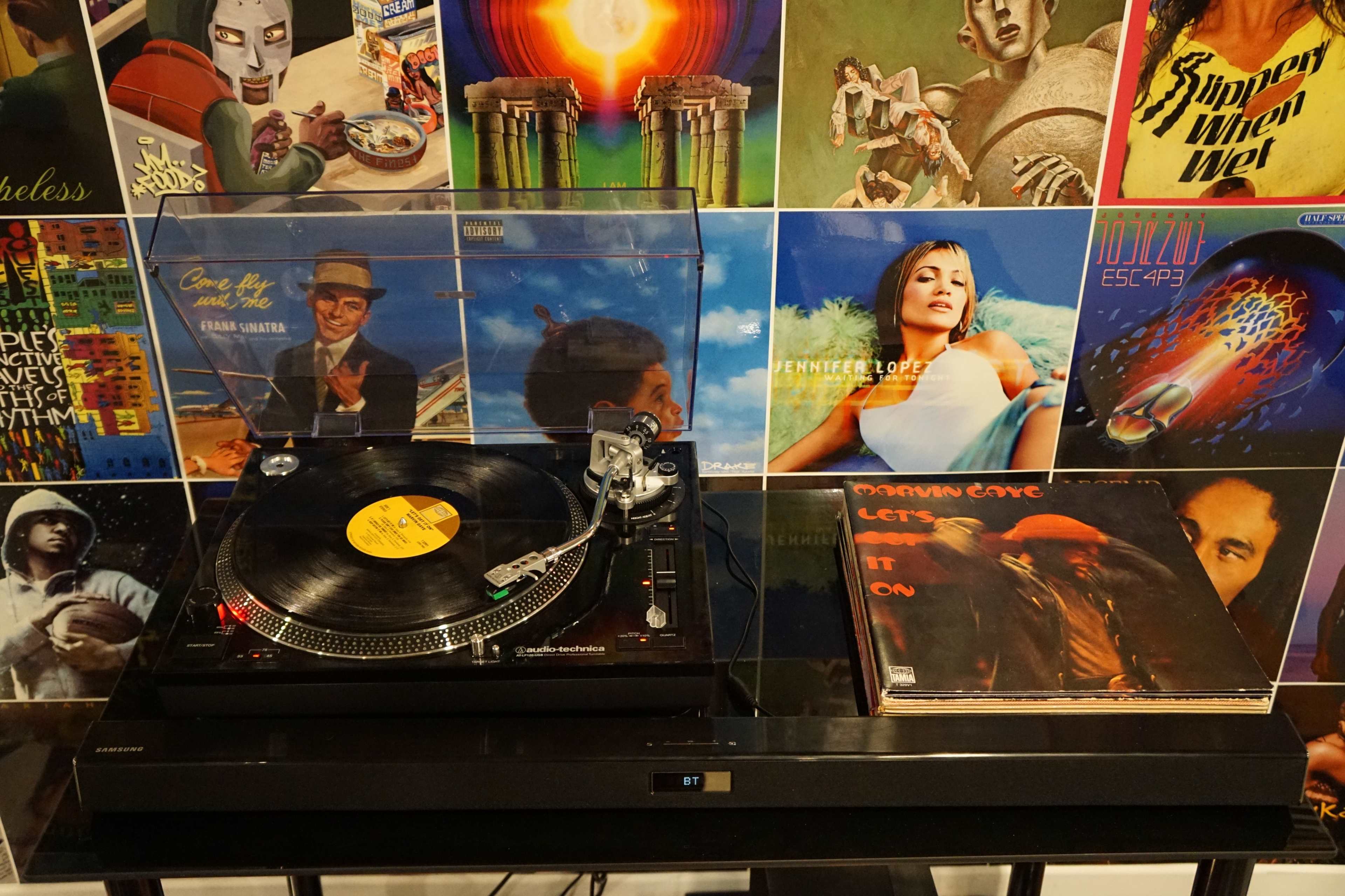 A turntable plays a vinyl record, surrounded by various album covers displayed on the wall behind it.