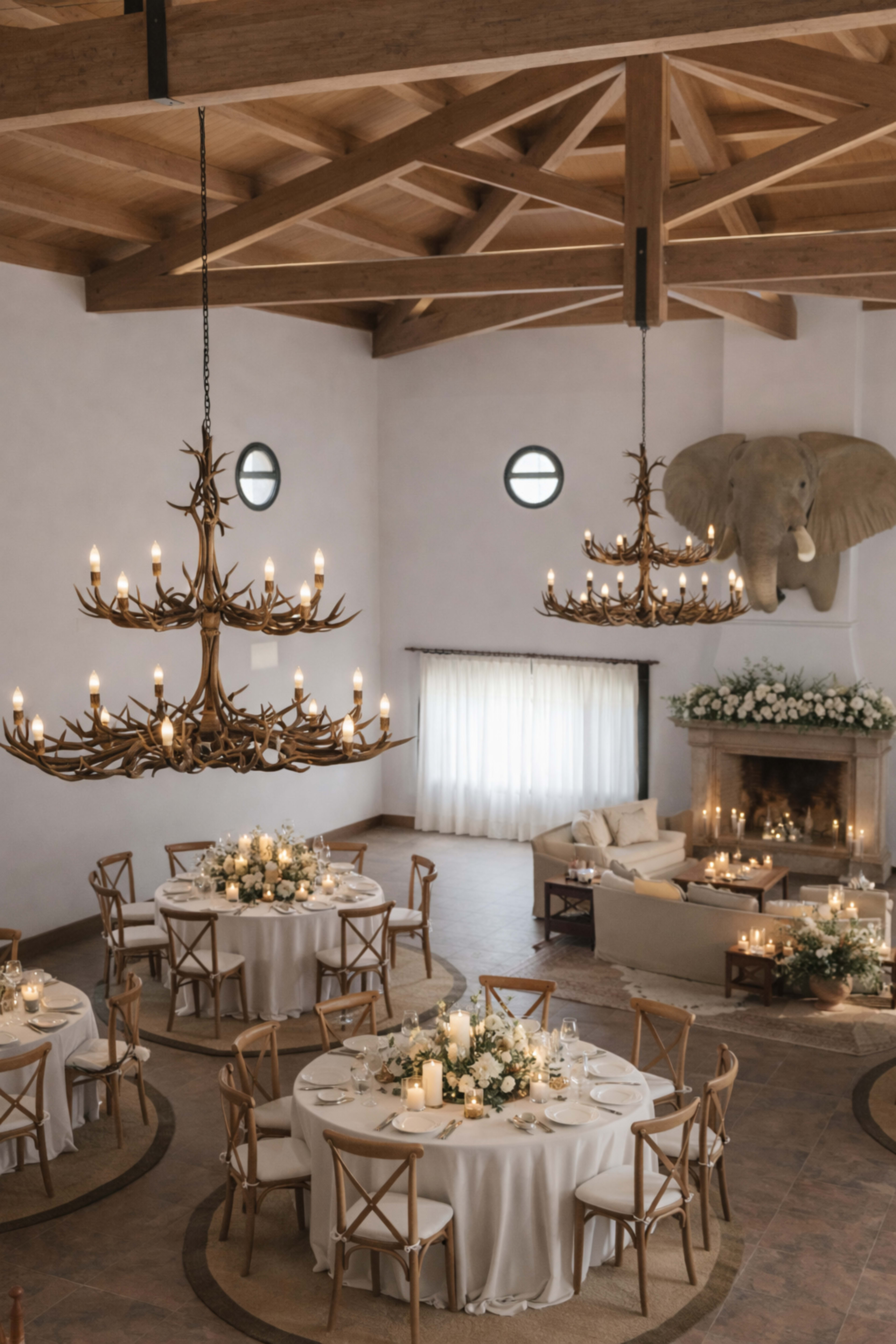 The image shows an elegantly decorated dining area with circular tables, candles, and floral arrangements, housed under a wooden beamed ceiling with large chandeliers and a mounted elephant head on the wall.
