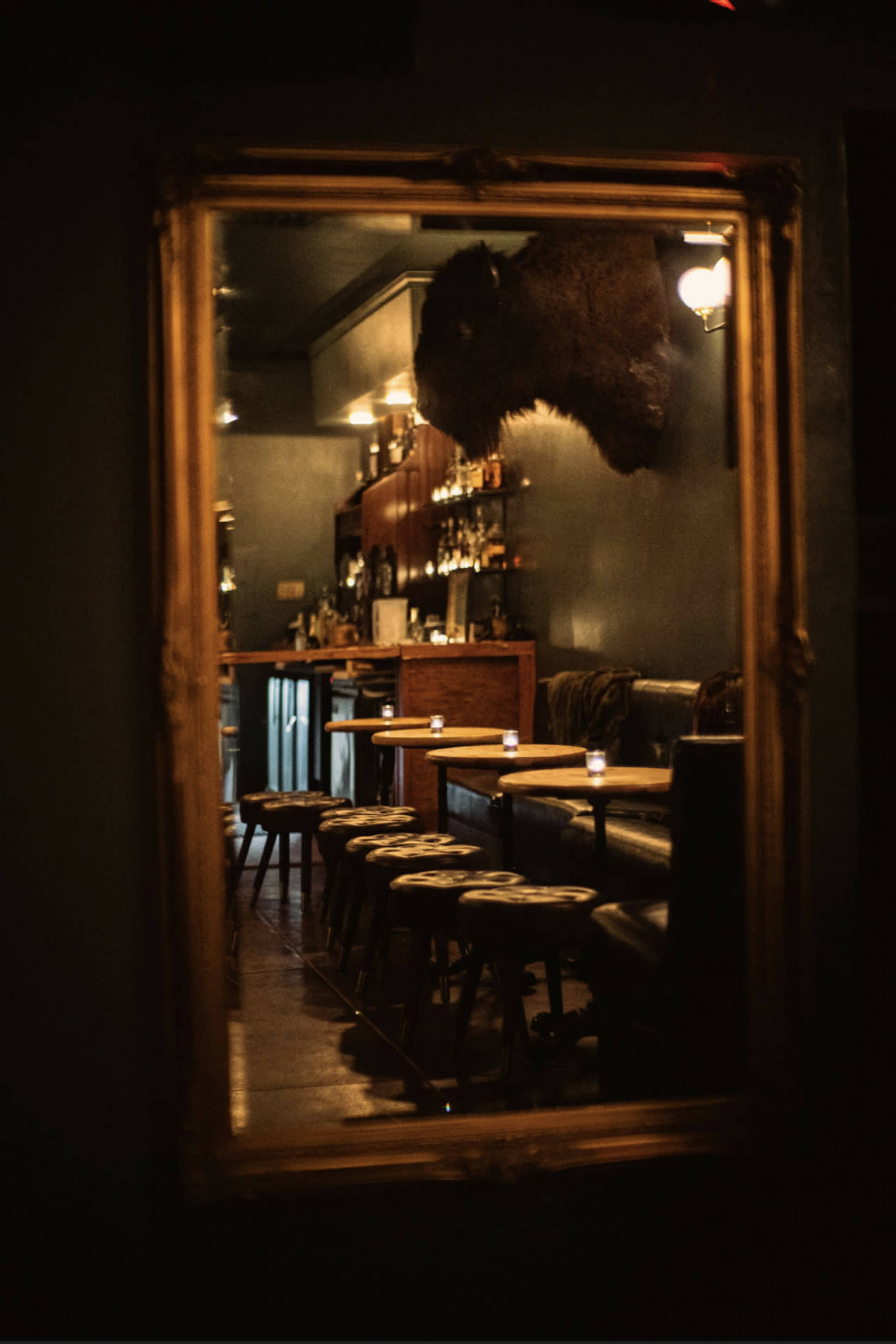 A bar area is reflected in a large ornate mirror, showcasing a mounted buffalo head on the wall and a row of stools in front of a dark countertop.