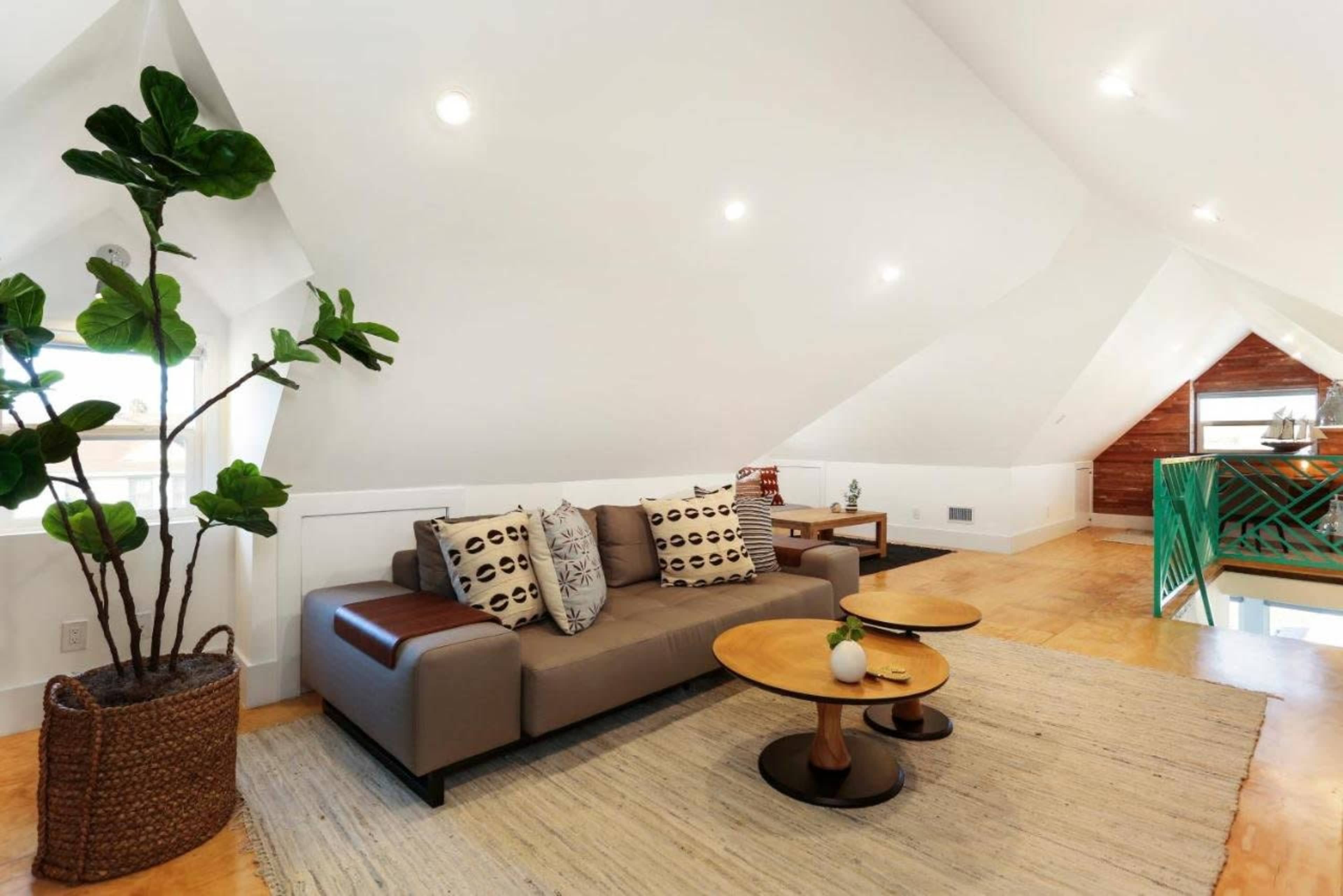 The image shows a cozy attic living area with a sofa, two round coffee tables, and a plant near a window under a slanted ceiling.