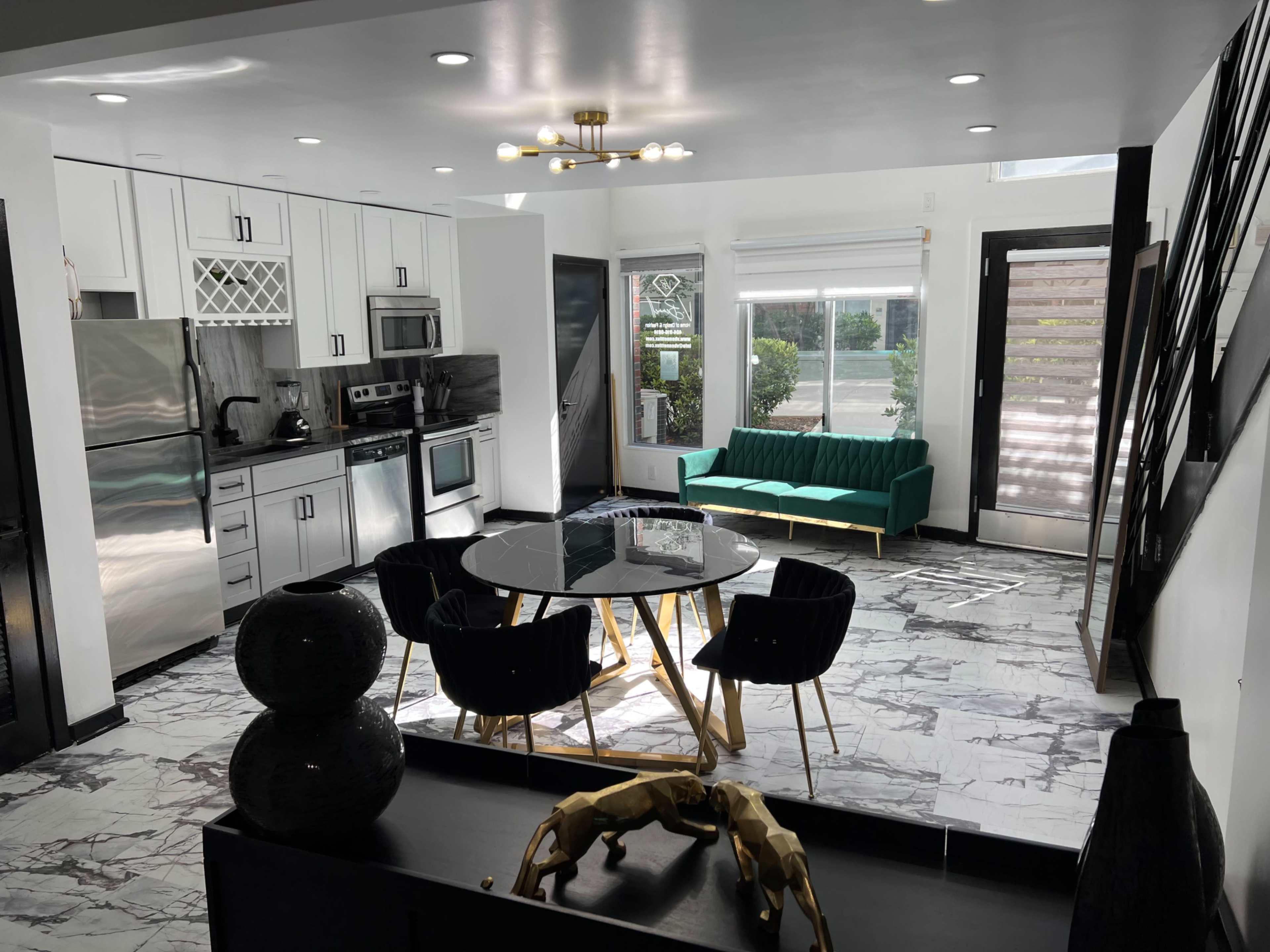 The image shows a modern kitchen and living area featuring a black and white marble floor, a green couch, a round dining table with black chairs, and stainless steel appliances.