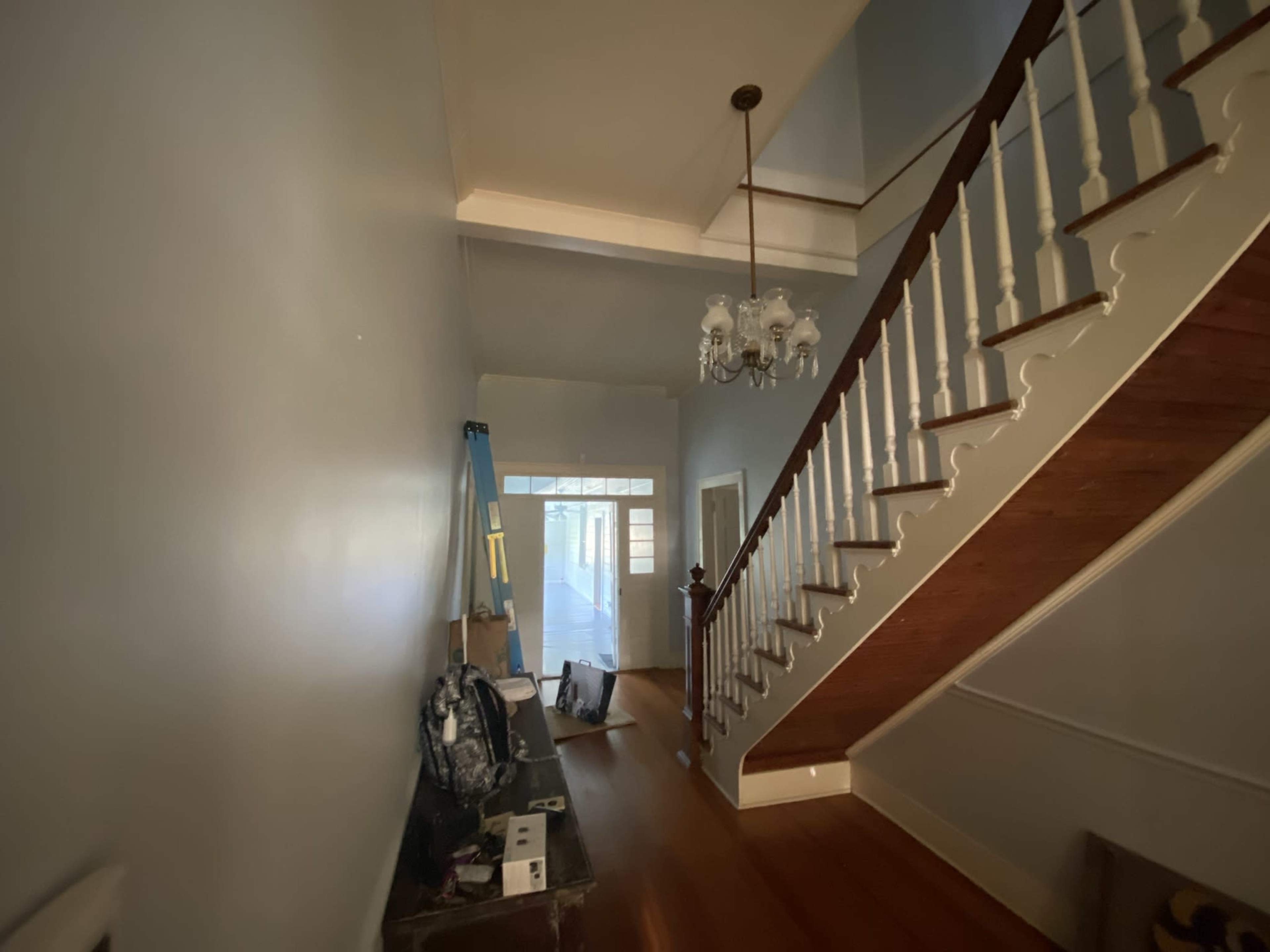 The image shows a hallway leading to a staircase, with a chandelier hanging from the ceiling and various items placed along the walls.