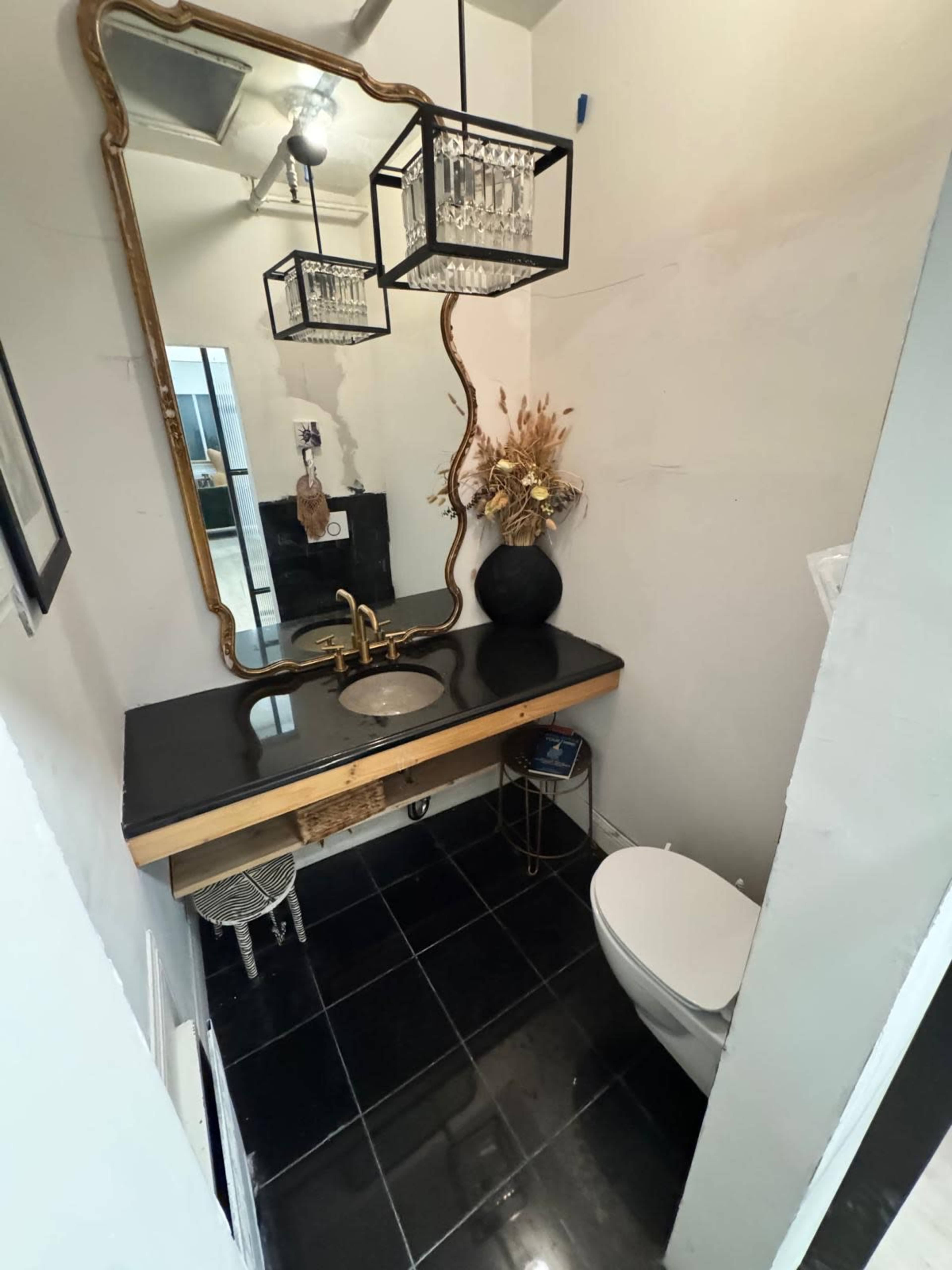 The image shows a small bathroom with a black countertop sink, a decorative mirror, a metal light fixture, and minimalist decor.