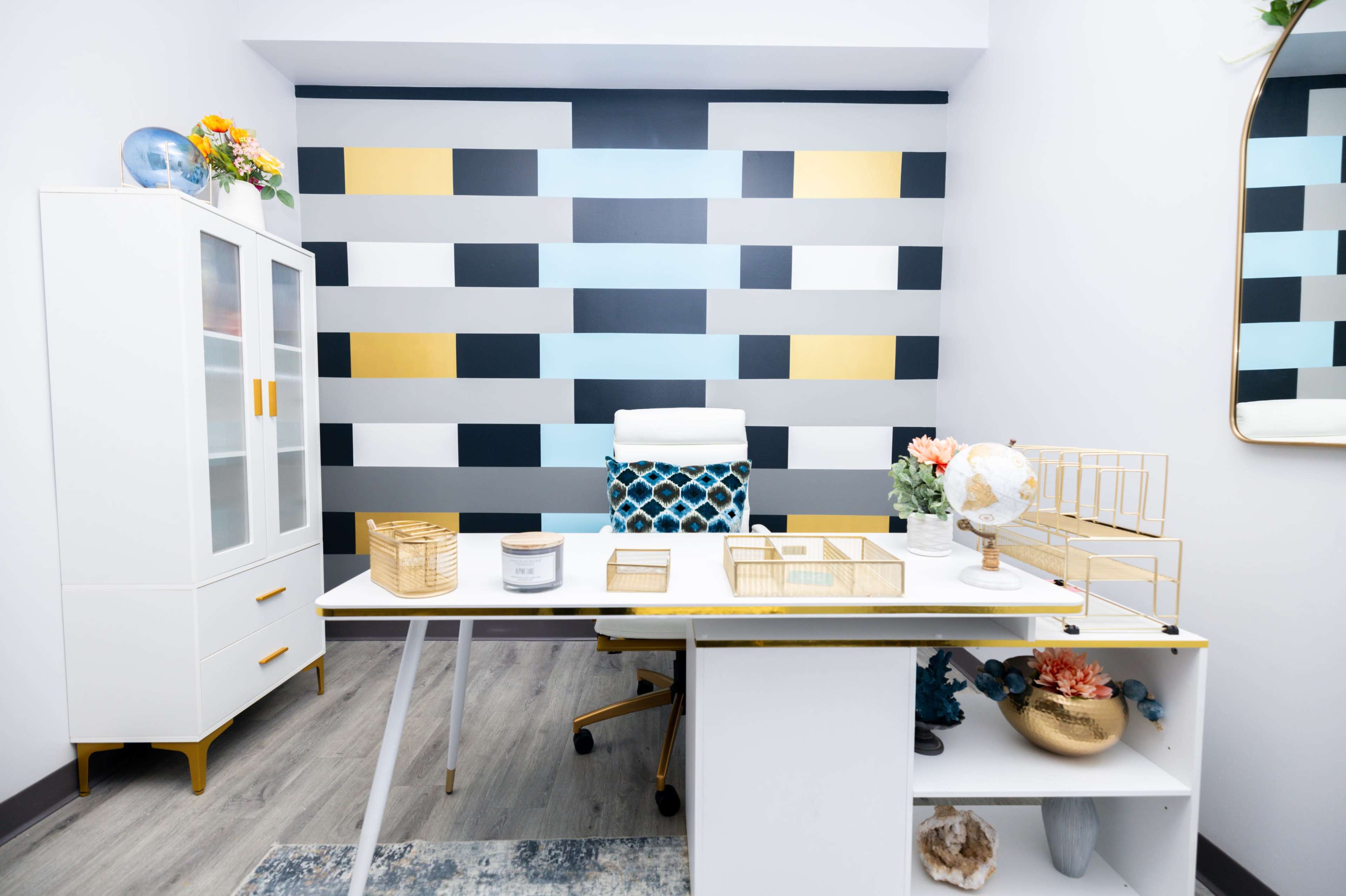 The image shows a modern office space featuring a white desk, a blue and gold accent chair, and a geometric wall design with various blocks of color.