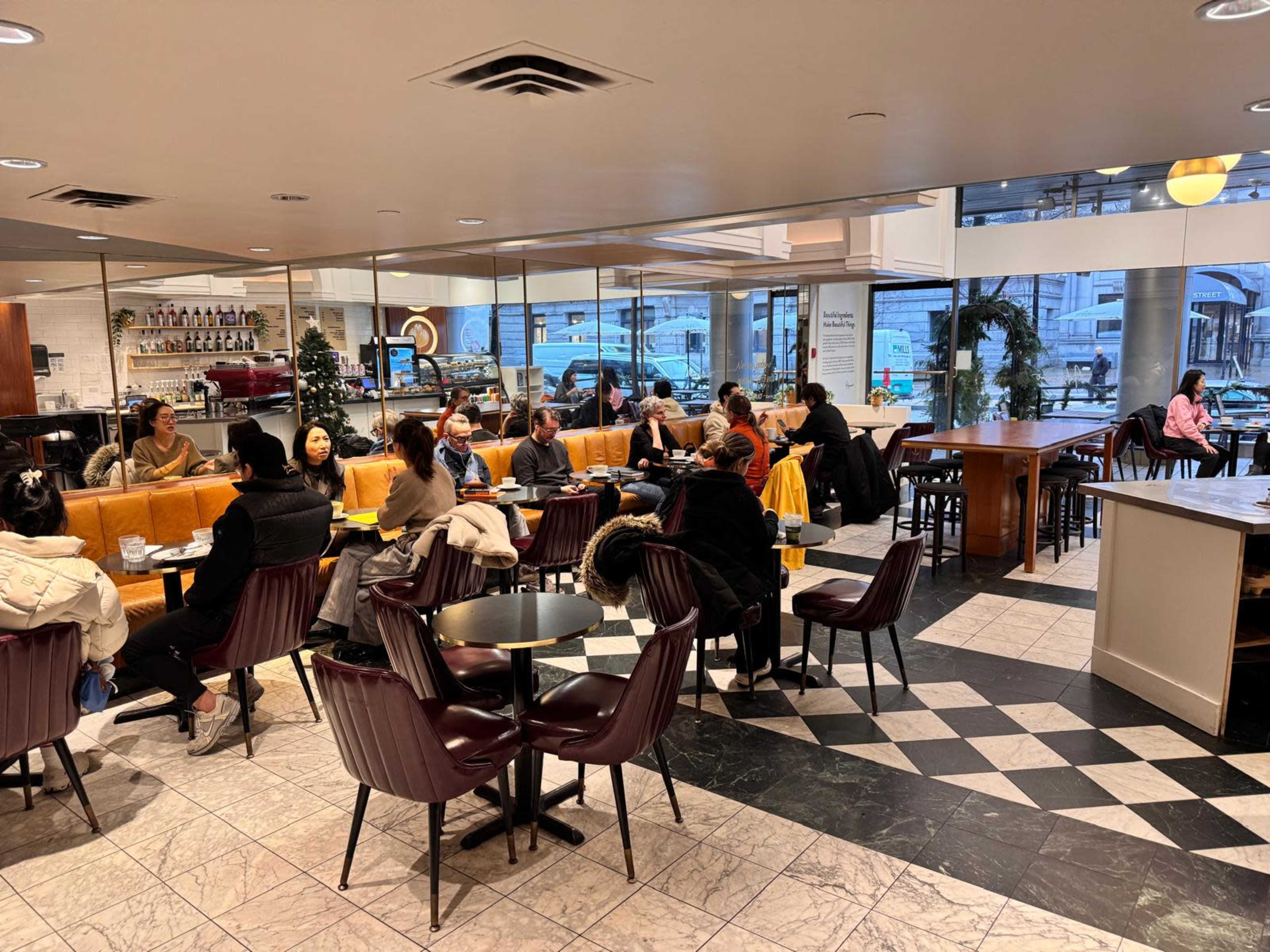 The image shows a busy café interior with several patrons seated at tables, some working on laptops, while large windows display a view of the street outside.