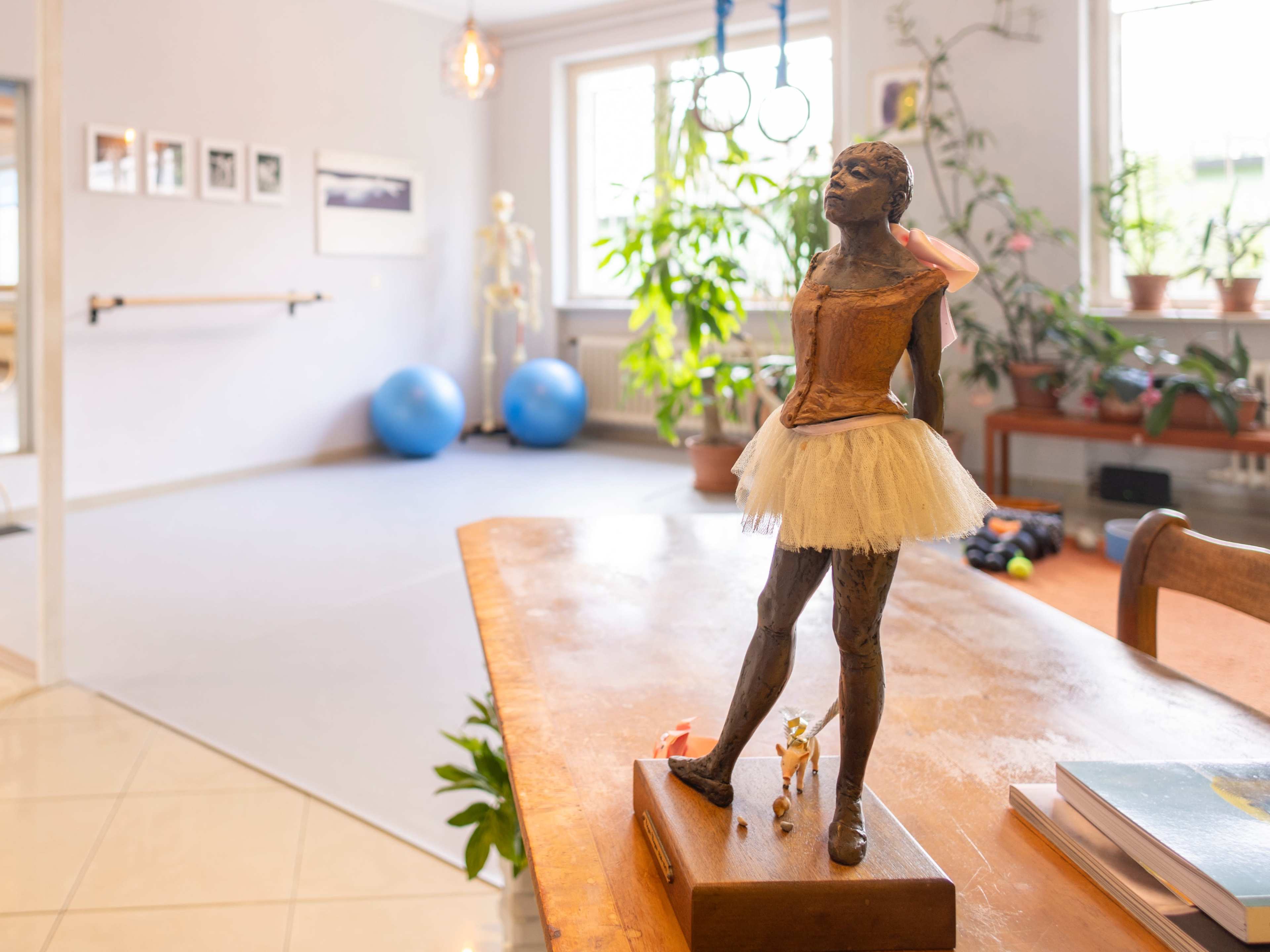 A ballet statue stands on a wooden table in a bright studio featuring exercise equipment and plants.