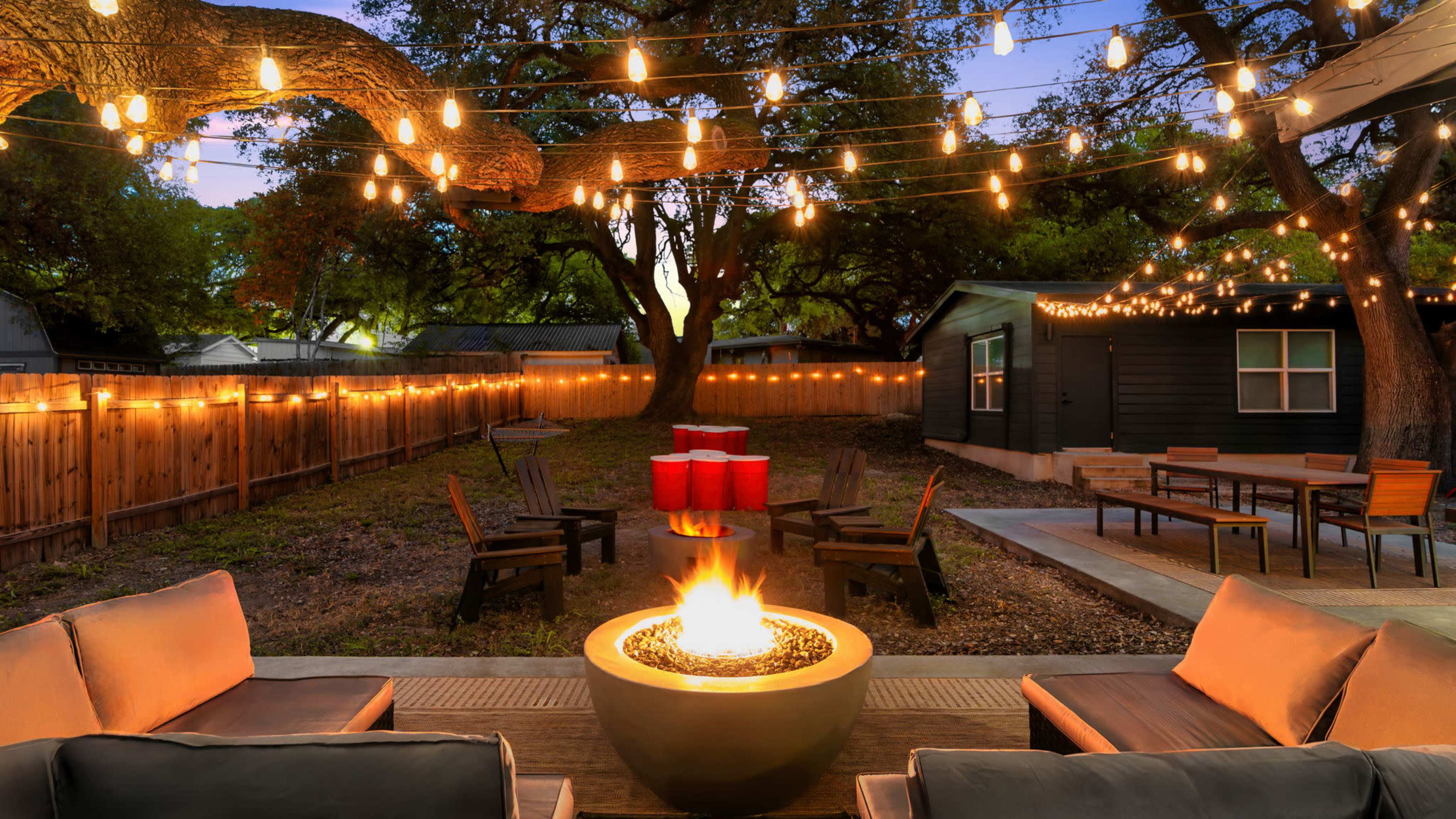 The image shows a backyard scene with a fire pit surrounded by seating, illuminated by string lights and a darkening sky.