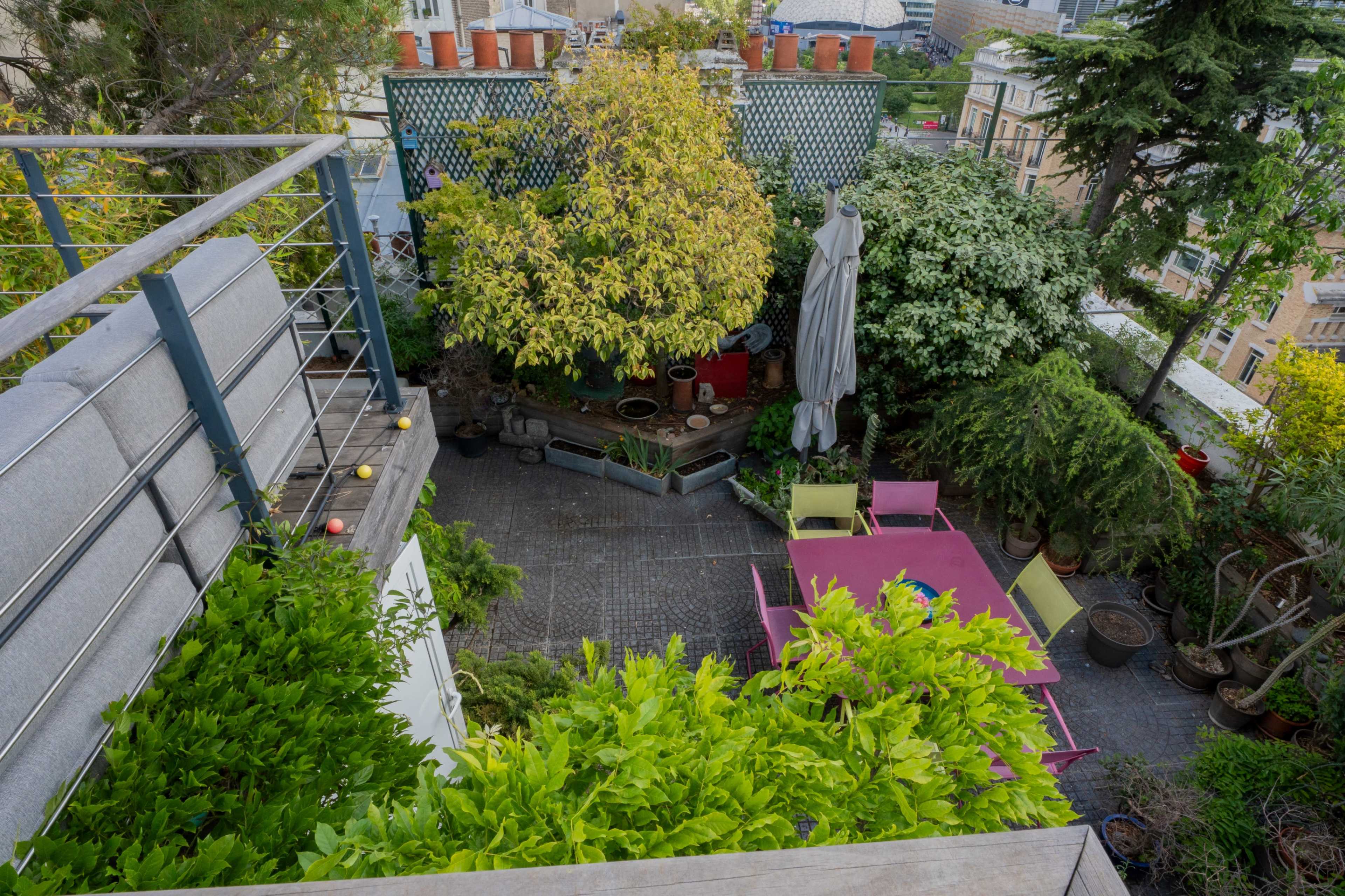 The image shows an aerial view of a green rooftop garden featuring various plants, a dining table with colorful chairs, and potted plants arranged throughout the space.
