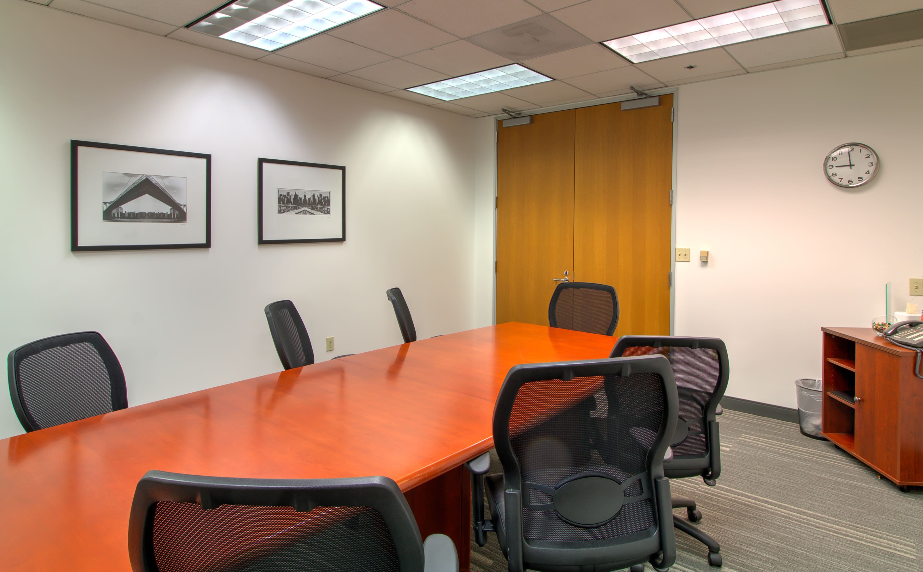 A conference room features a long wooden table surrounded by black ergonomic chairs, with framed black-and-white photographs on the walls and a clock above a side table.