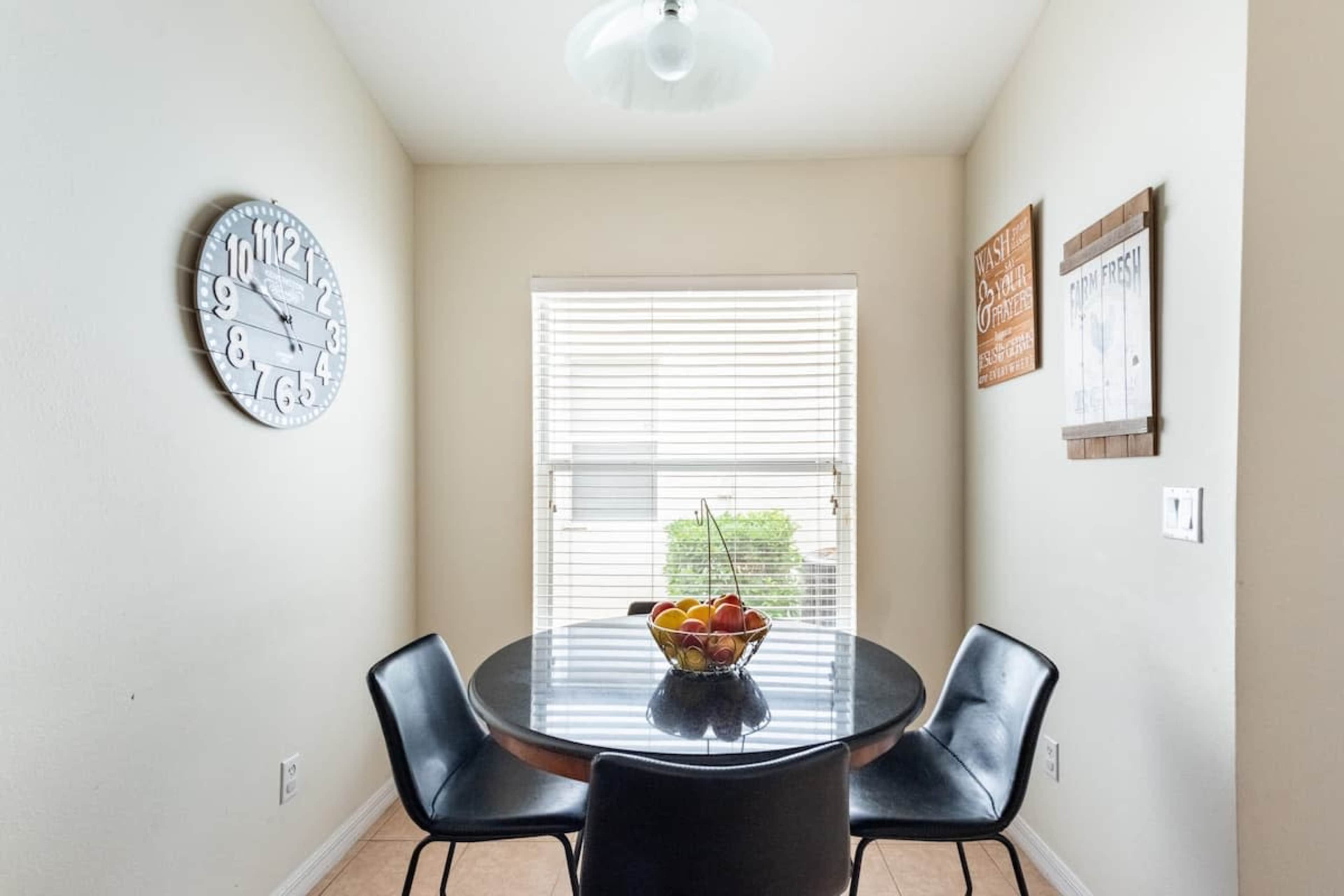 A small dining area features a round black table with a fruit bowl and four black chairs, set against a backdrop of light-colored walls and a window with blinds.