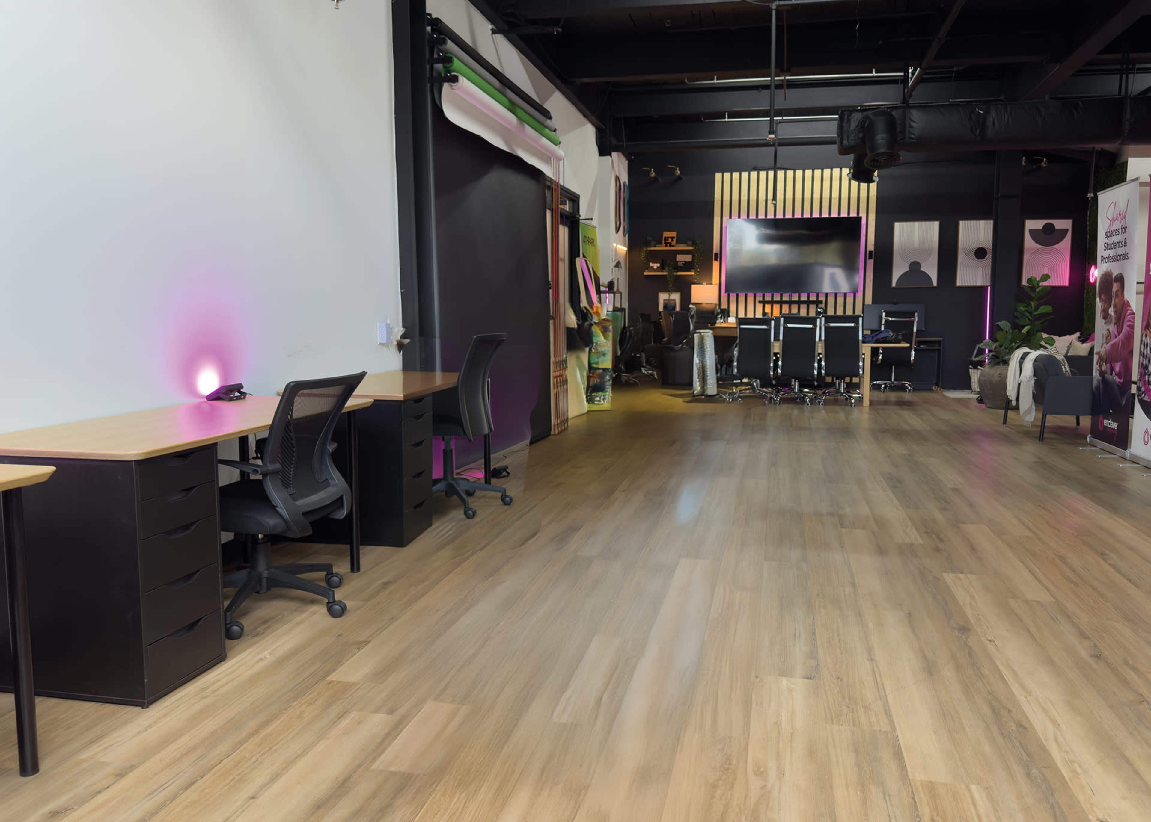 The image shows a modern office space featuring empty desks on one side and a conference area with chairs and a large screen on the other.