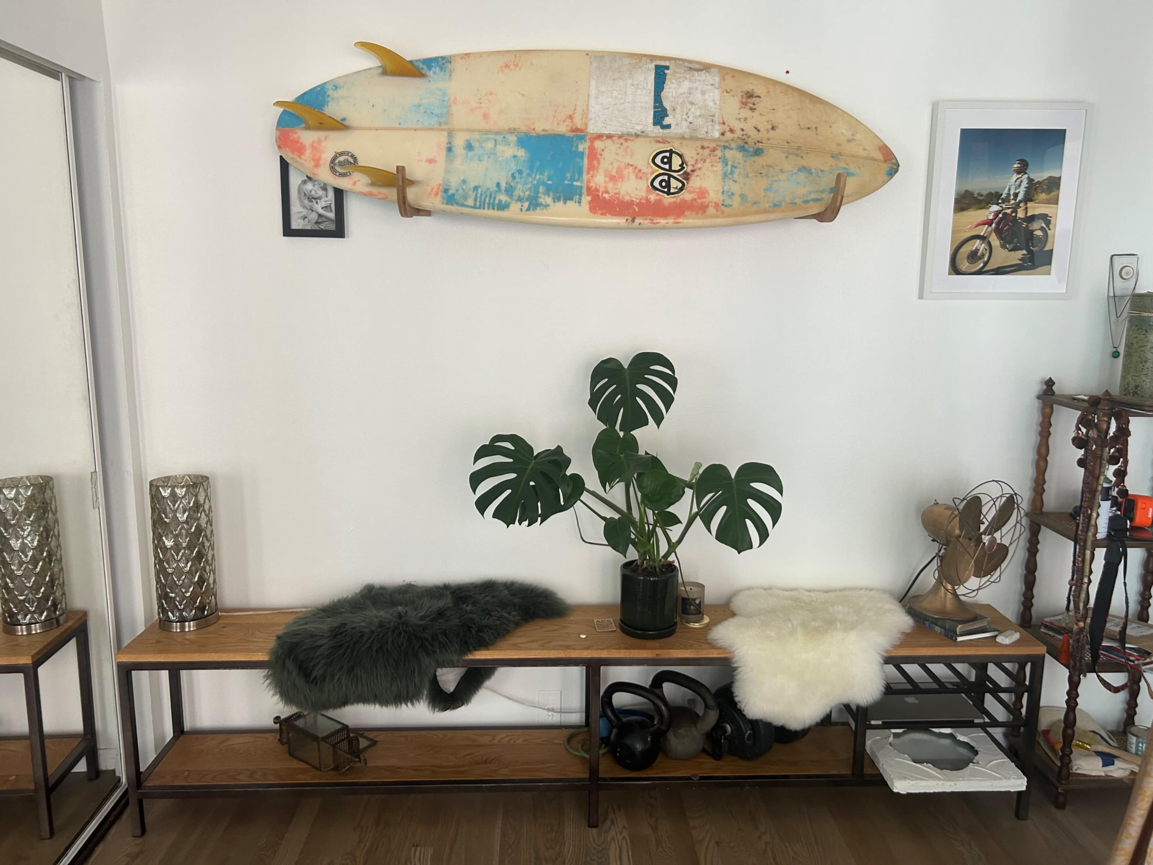 A wooden bench with decorative throws and a potted plant is positioned beneath a surfboard mounted on the wall alongside a framed photo.