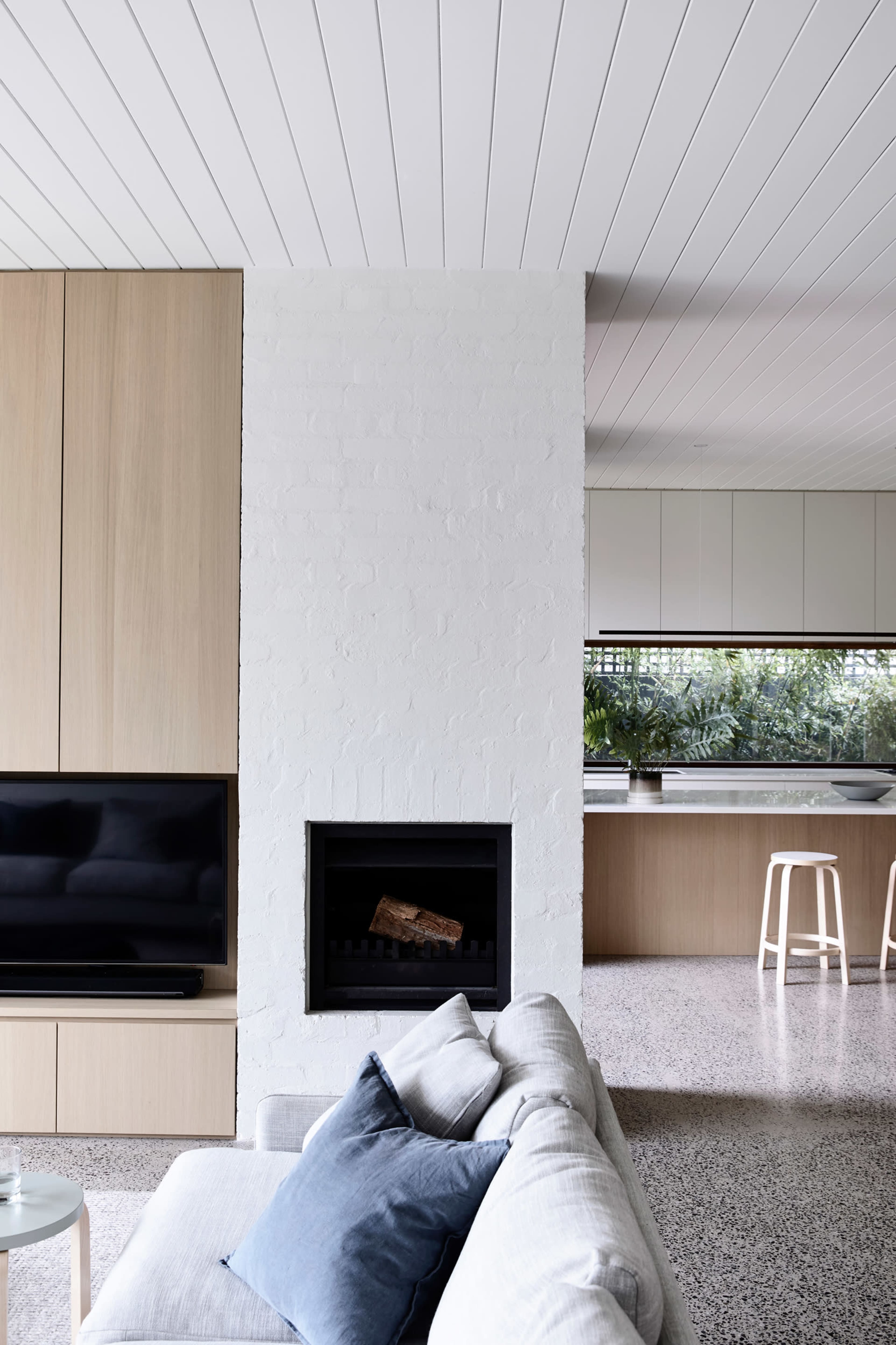 A modern living room featuring a light-colored sofa, a wall-mounted television, and a white brick fireplace, with a kitchen area visible in the background.
