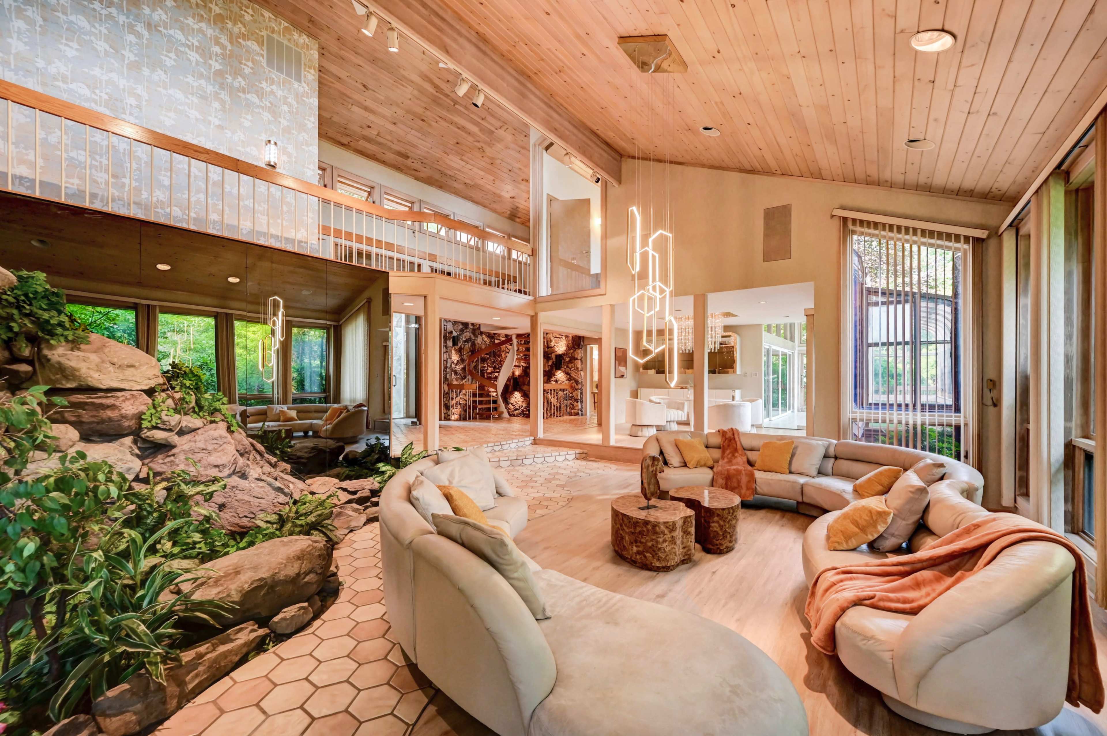 The image shows a spacious living room with a circular sofa, wooden ceiling, large windows, and natural stone elements integrated into the design.