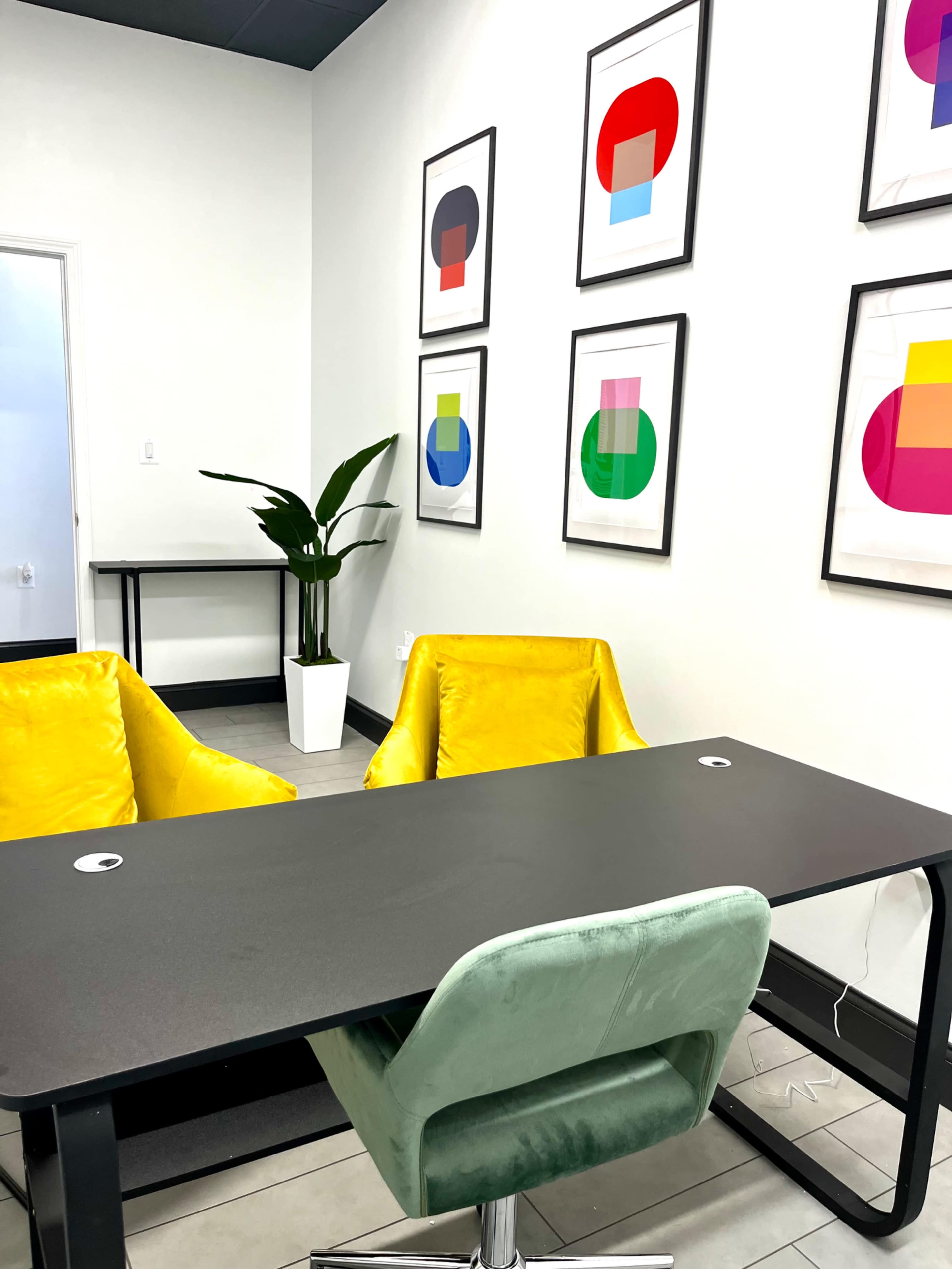 The image shows a modern office with two yellow chairs and a green office chair facing a black desk, while colorful abstract art pieces hang on the wall.