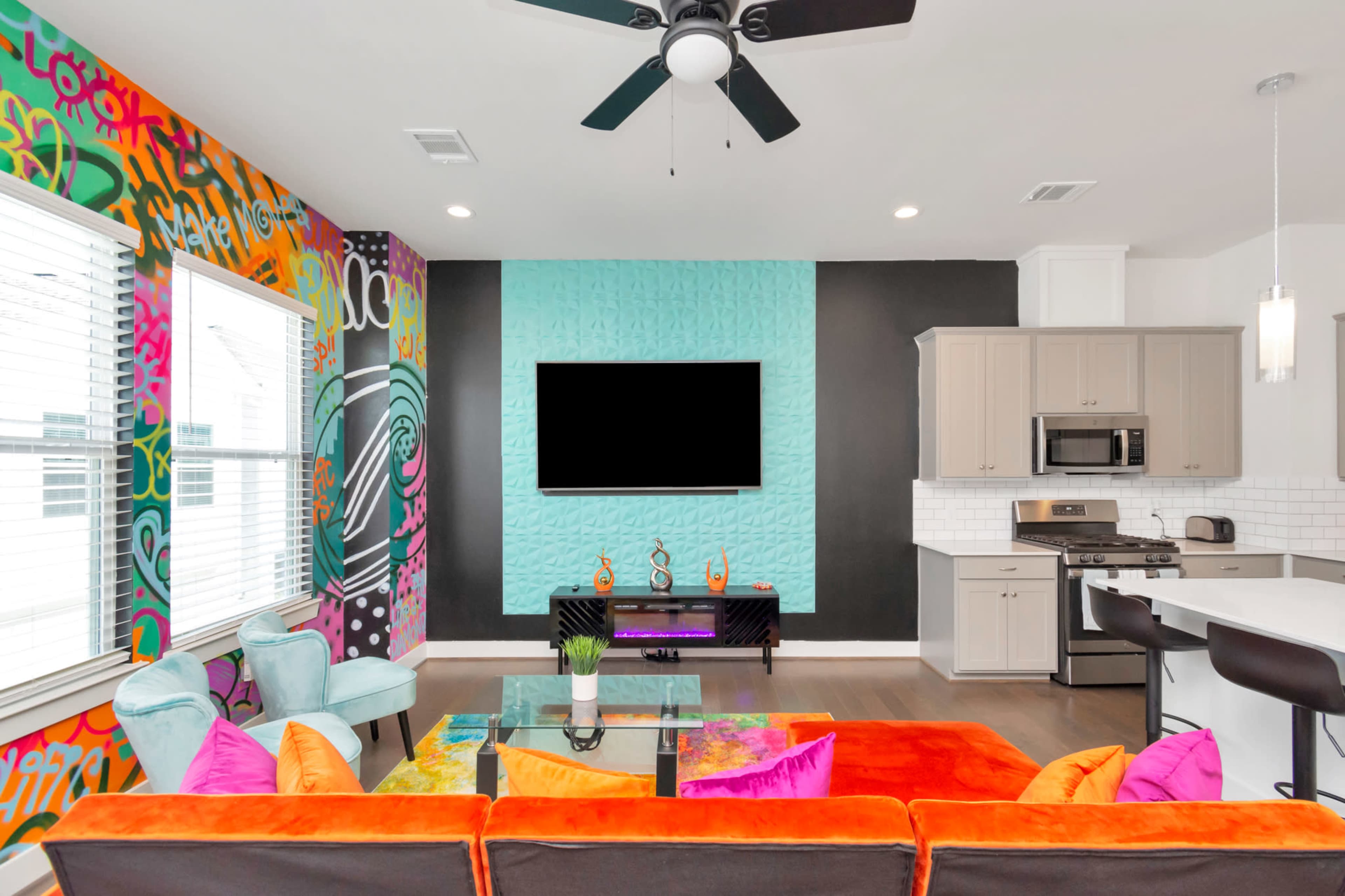 The image shows a modern living room with colorful wall art, a large television mounted on the wall, and a sectional sofa in vibrant colors.