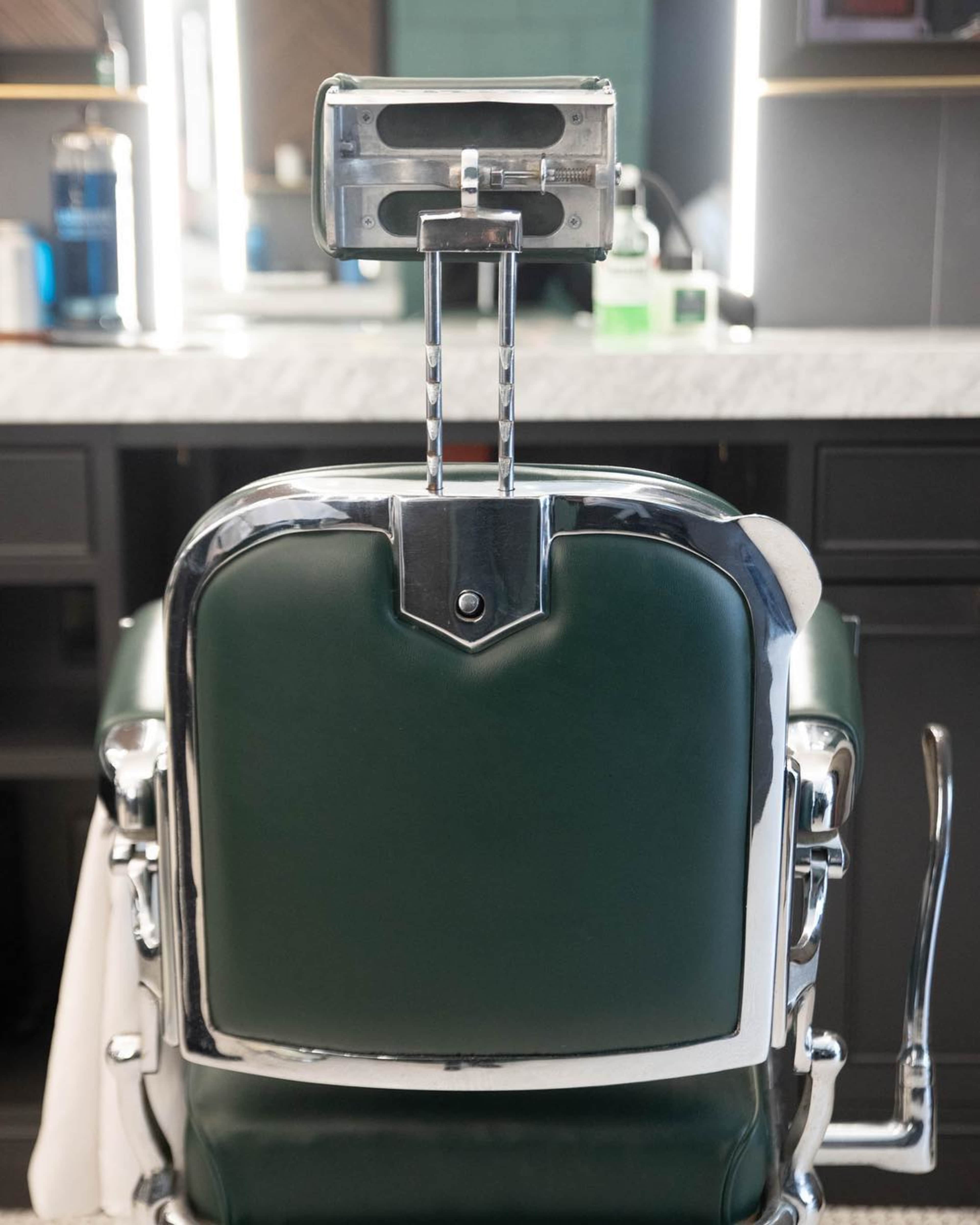 A green leather barber chair with chrome accents is positioned in front of a mirror and countertop in a barbershop.
