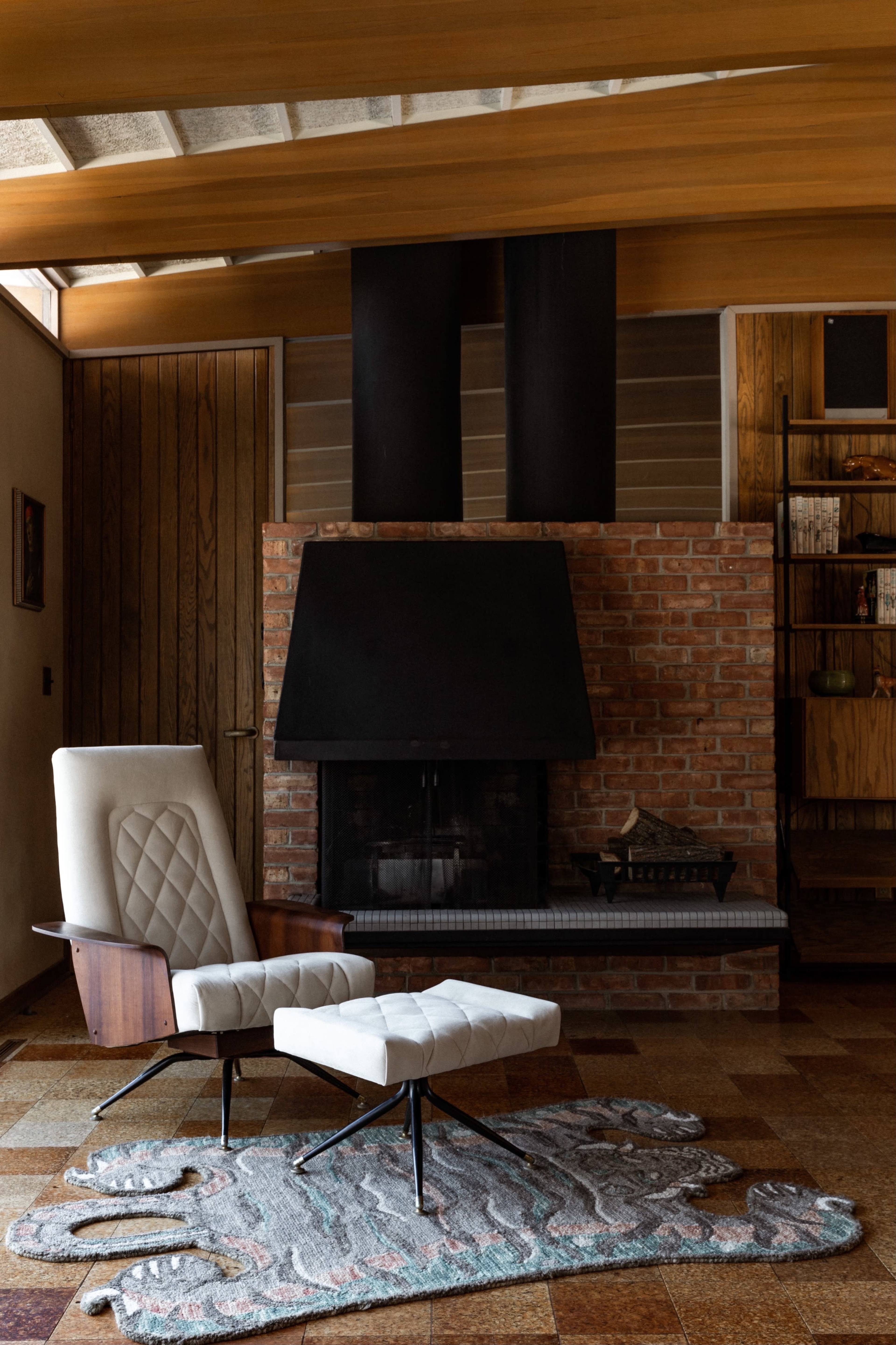 The image shows a modern lounge area featuring a white upholstered chair and ottoman in front of a brick fireplace, with wooden beams and a patterned rug on the floor.
