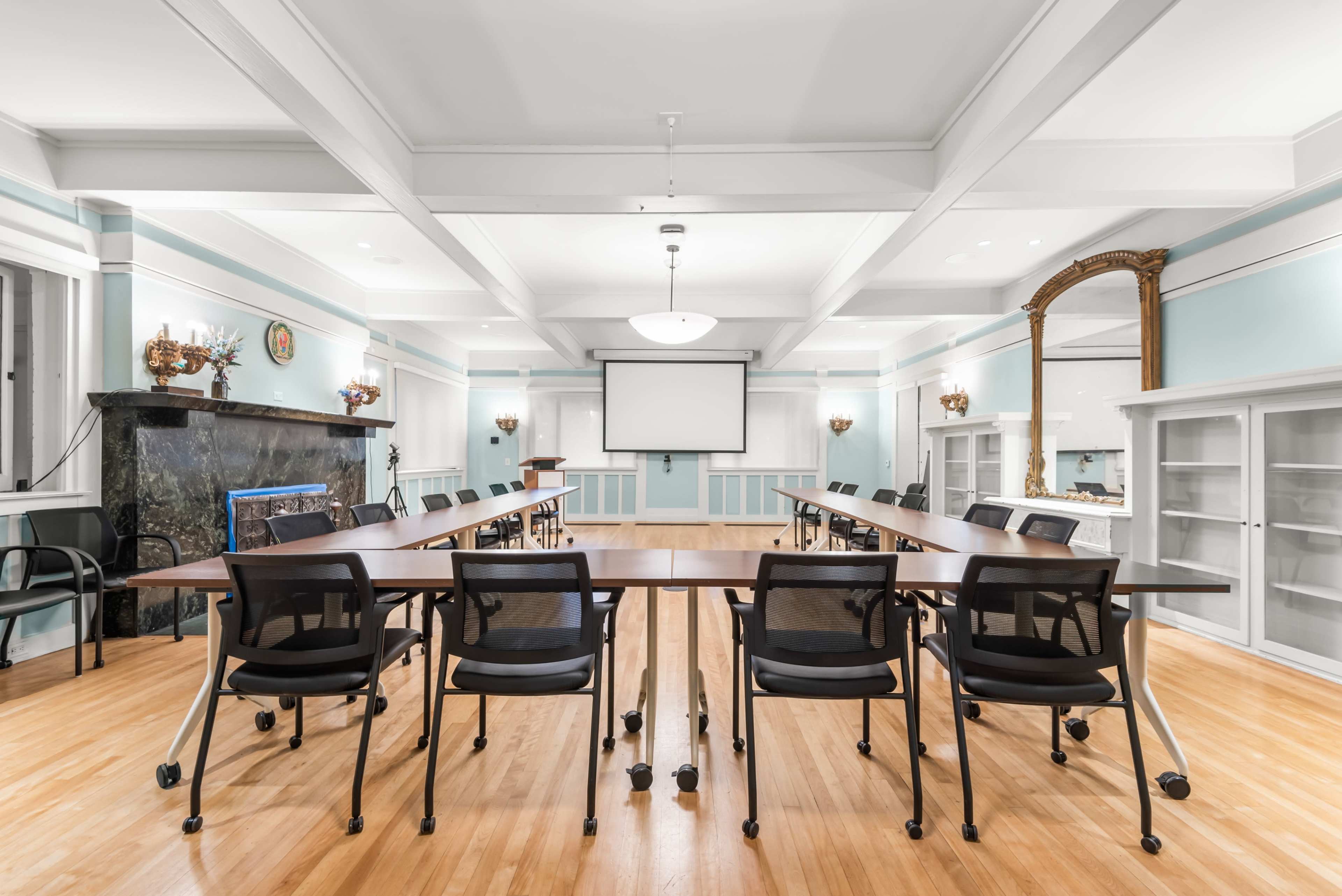 A spacious conference room features a long, rectangular table surrounded by rolling chairs, with a screen at one end and light blue walls adorned with decorative elements.