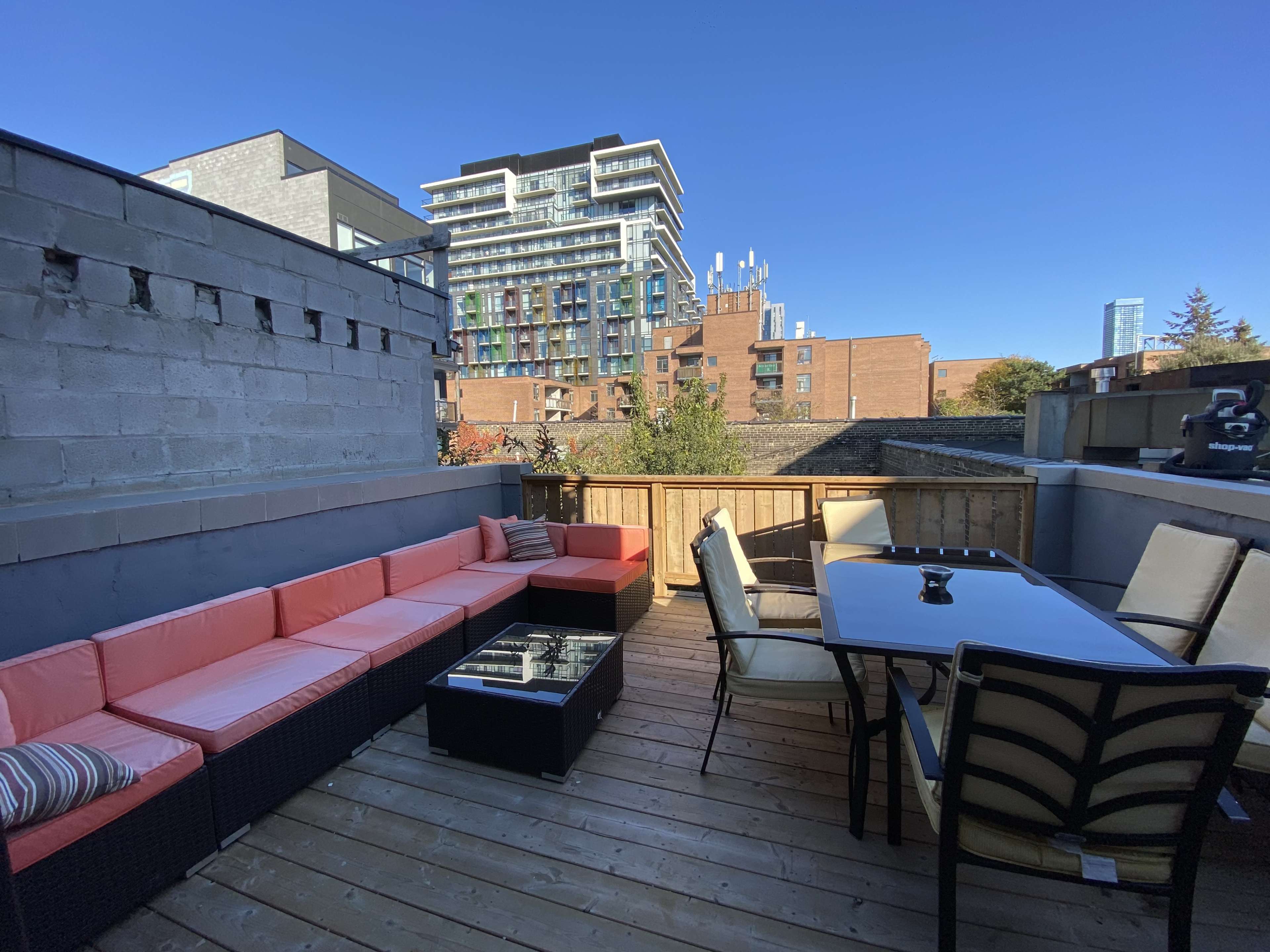 A balcony with a wooden deck featuring a seating area with orange cushions, a glass-topped table, and a view of buildings in the background.