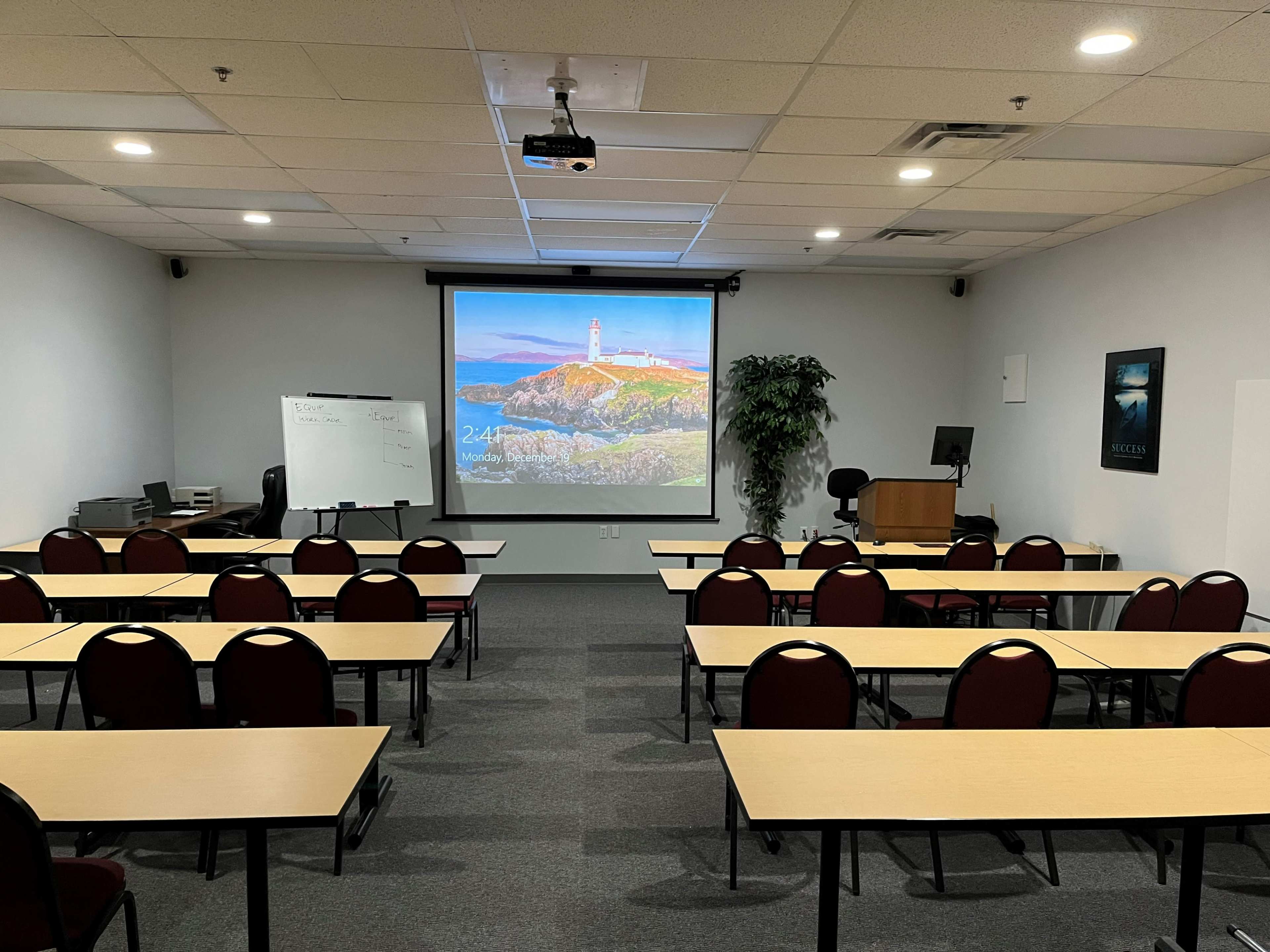A classroom setting with rows of tables and chairs facing a large projection screen displaying a coastal image.