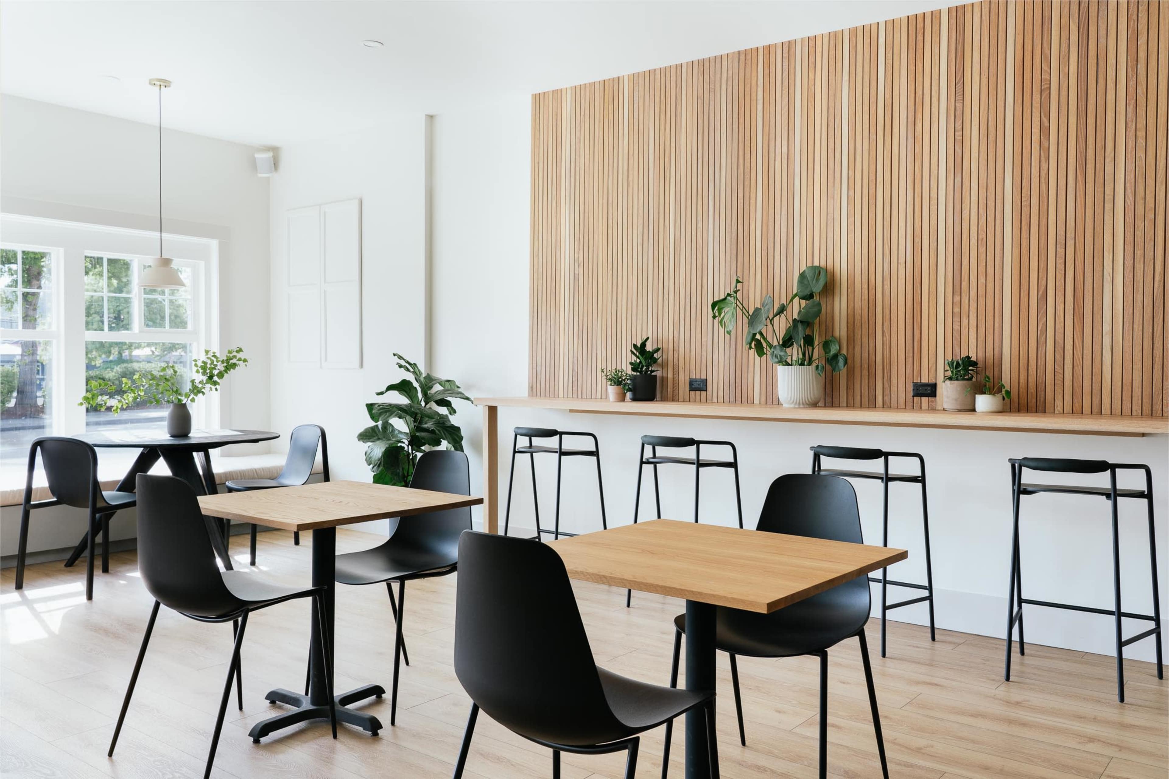 A modern café interior with wooden walls, minimalist furniture, and plants arranged on tables and a counter.