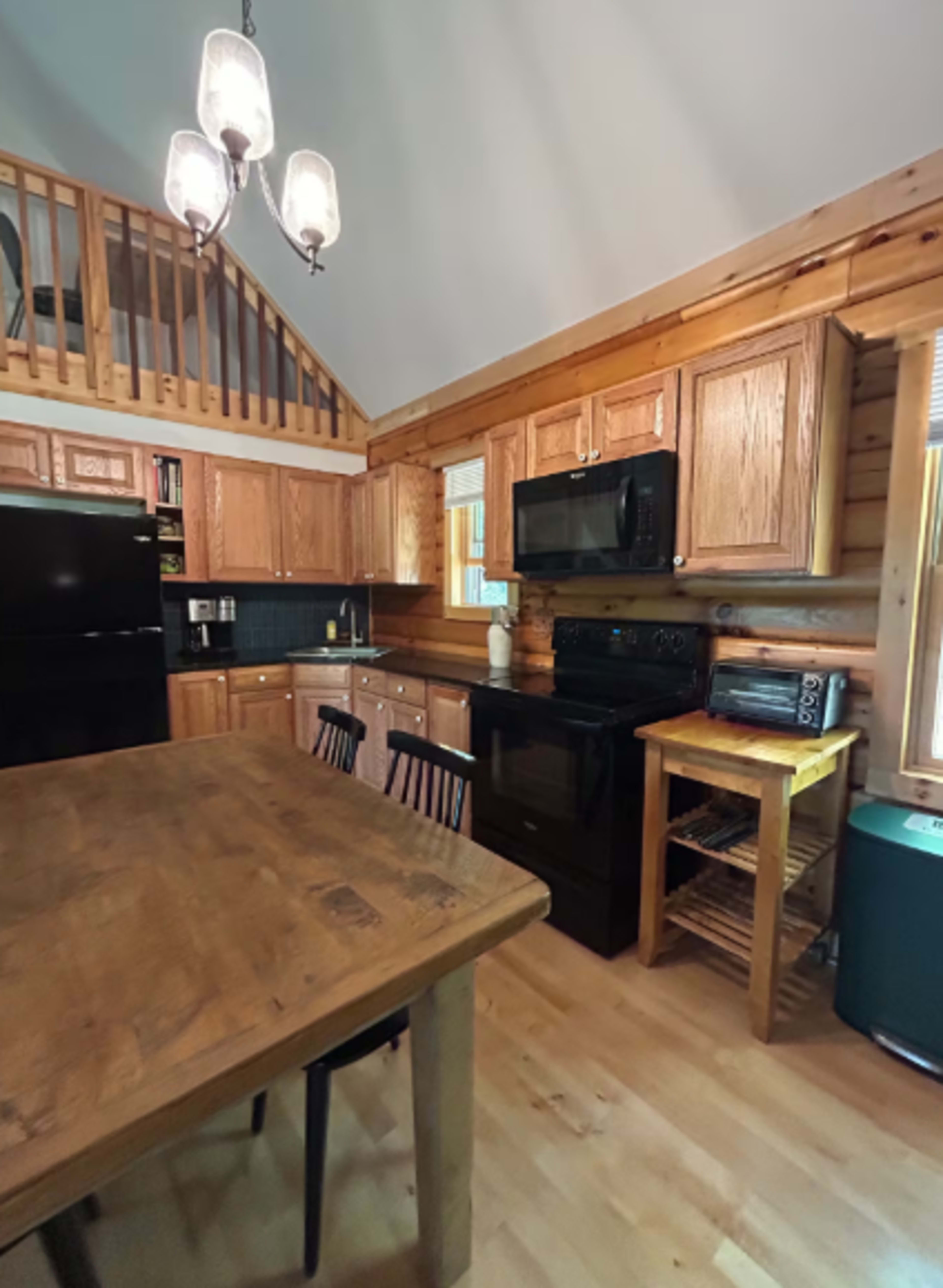 The image shows a wooden cabin kitchen with a dining table, black appliances, and rustic cabinetry.