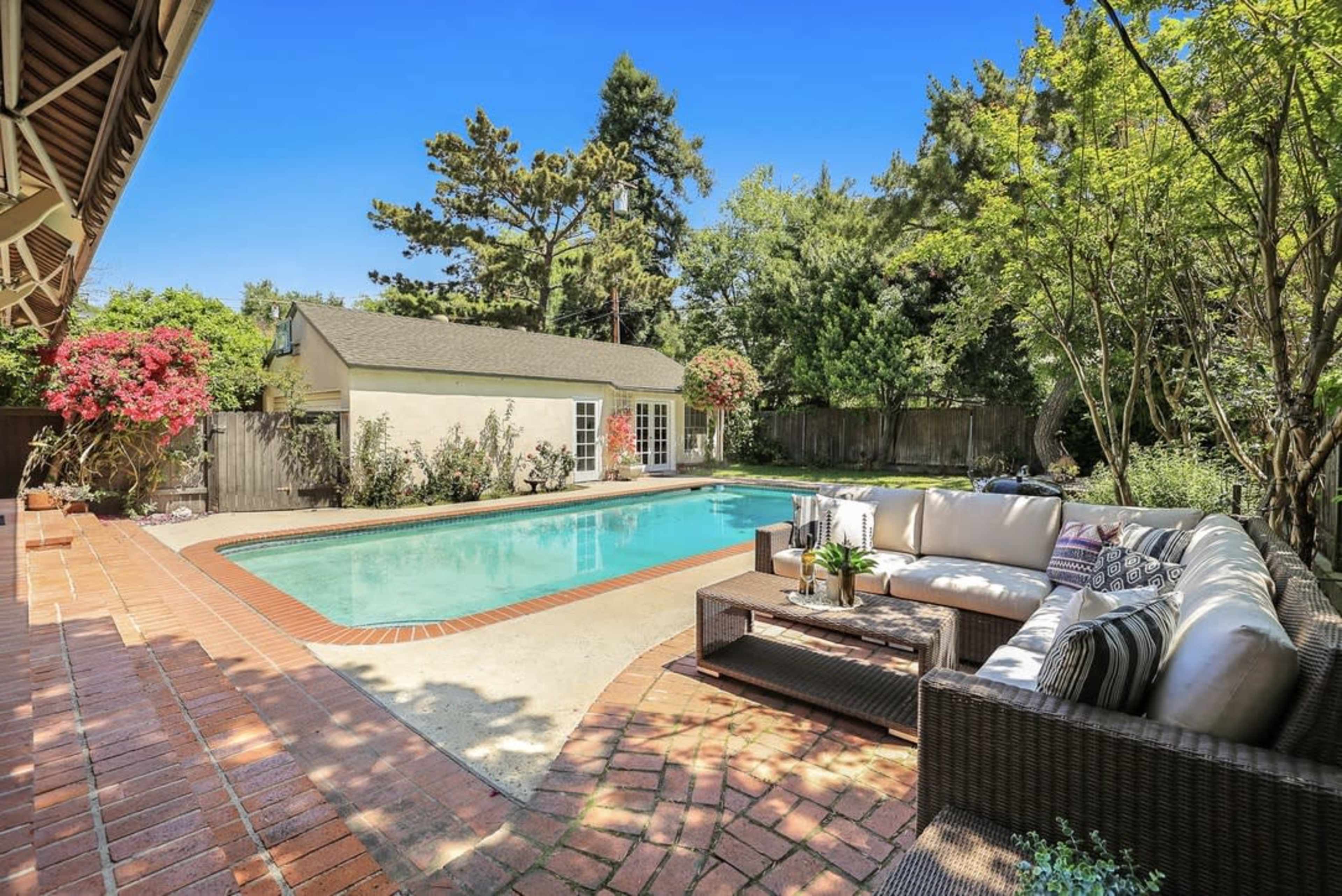 A backyard features a rectangular pool surrounded by a brick patio and a seating area with a sofa, overlooking a well-maintained garden.