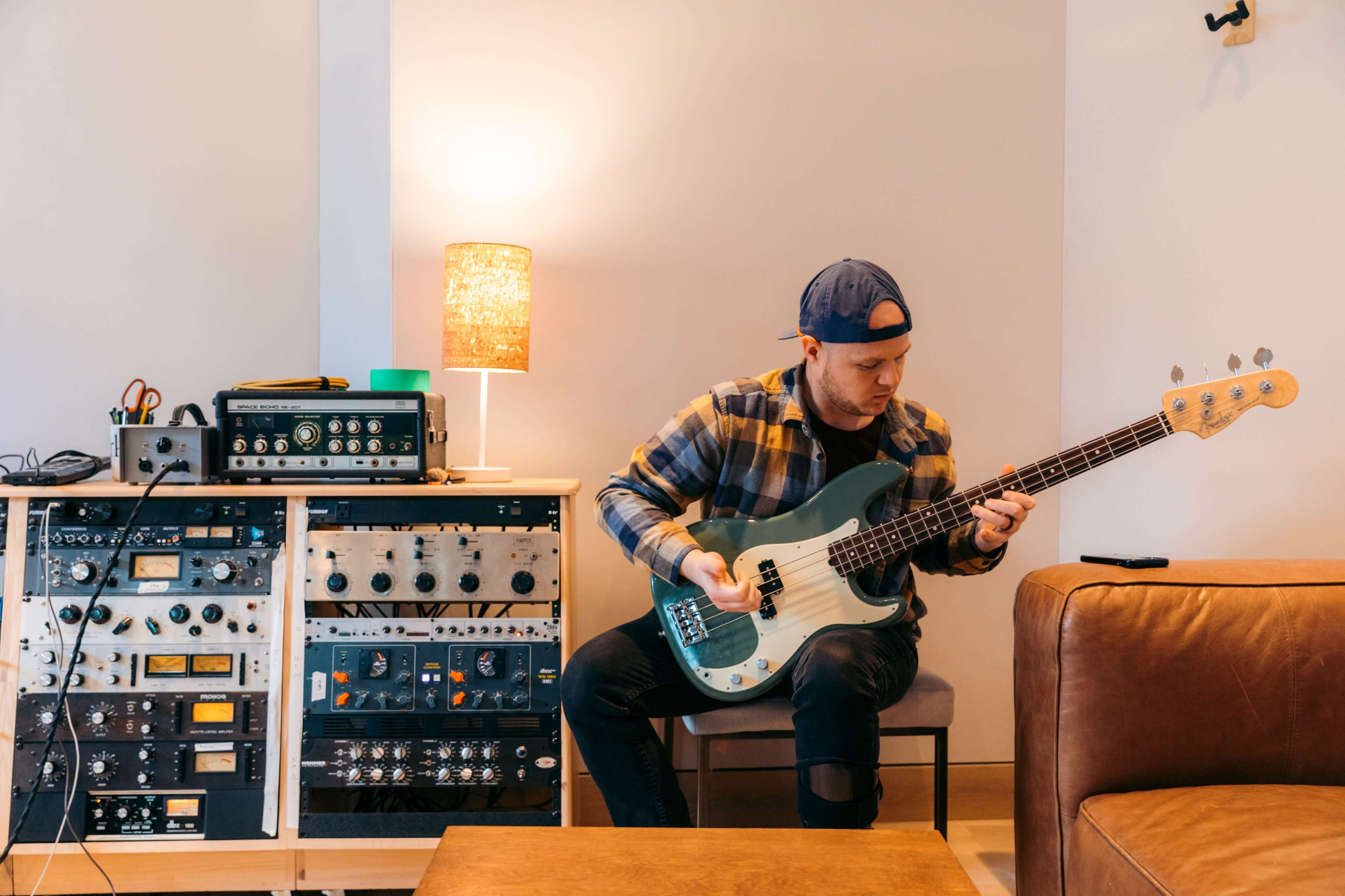 A man in a cap plays a bass guitar while sitting on a chair beside a console of audio equipment and a lamp.