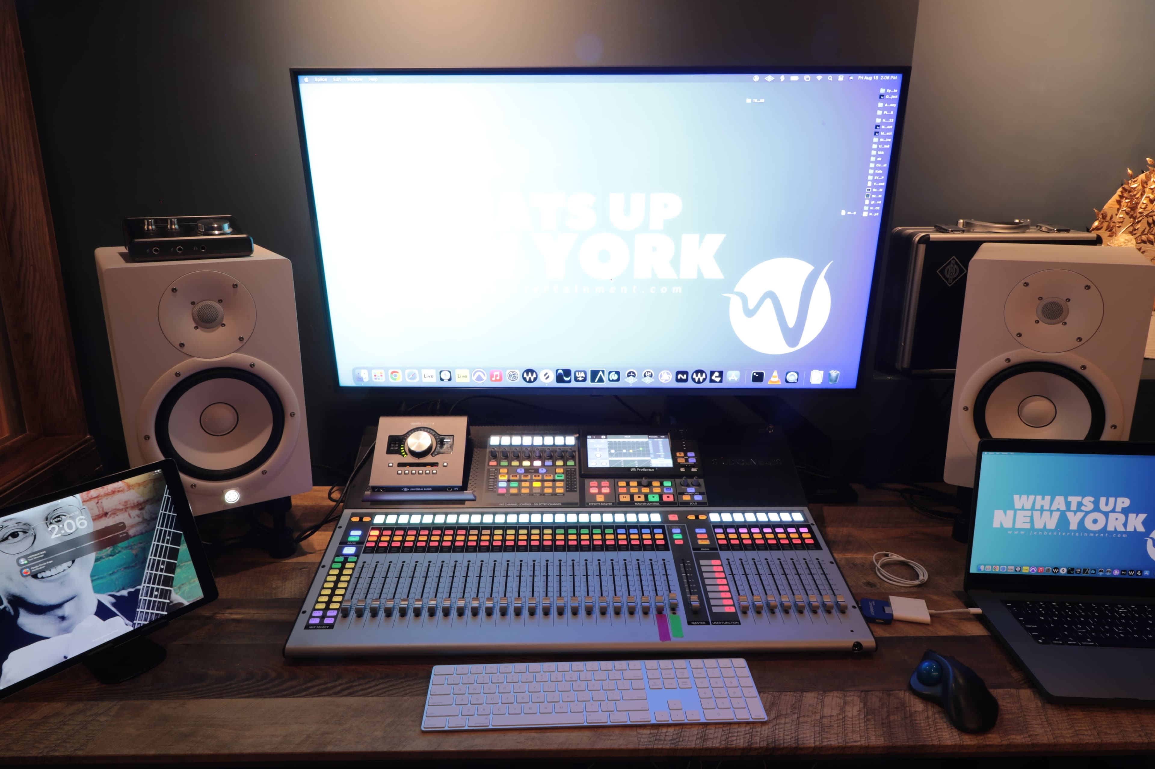 A digital audio workstation setup features a large monitor displaying a logo, a control surface with colorful buttons, and two studio monitors, alongside a laptop and tablet on a wooden desk.