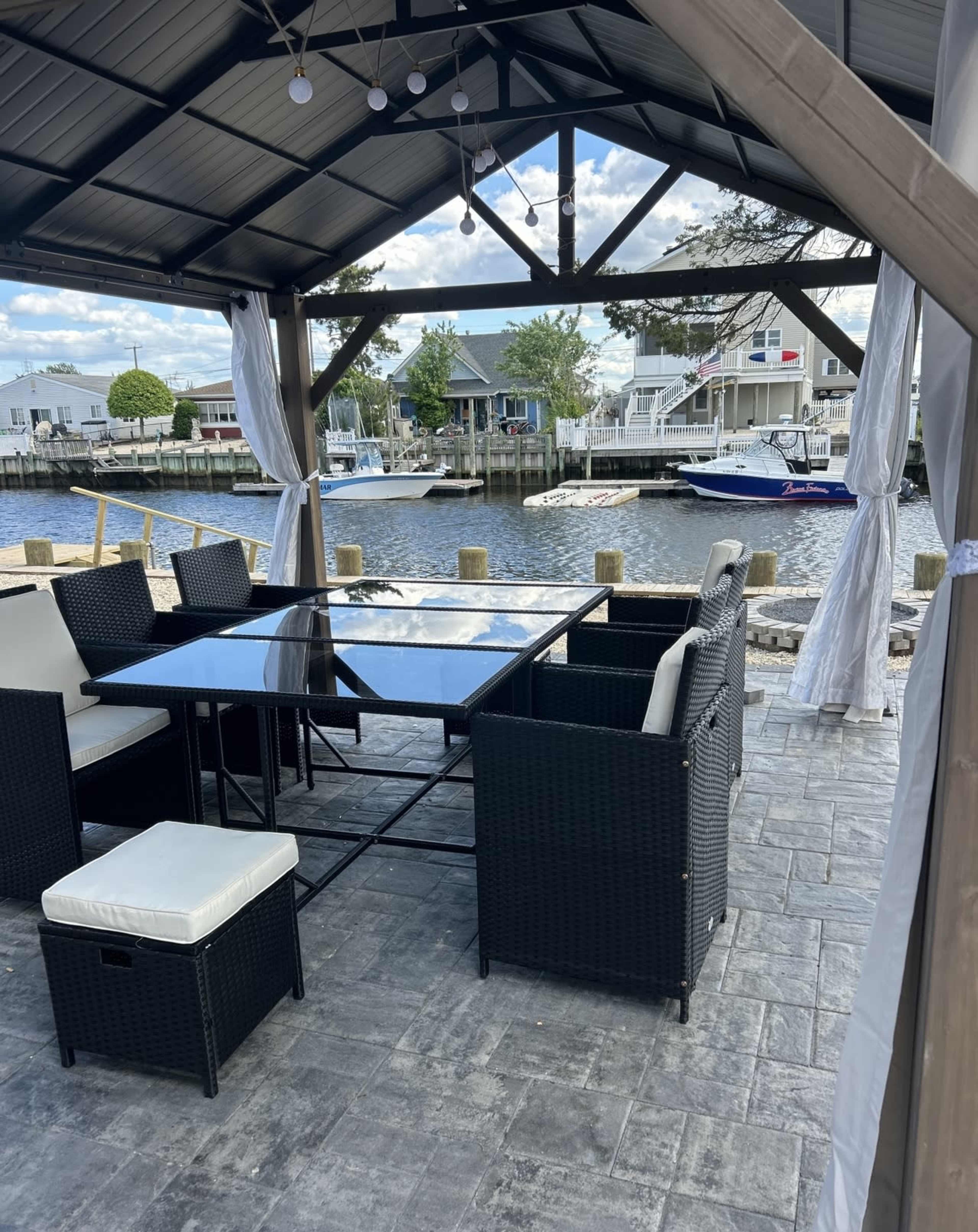 A covered outdoor seating area with a glass table and rattan chairs overlooks a canal lined with boats and houses.