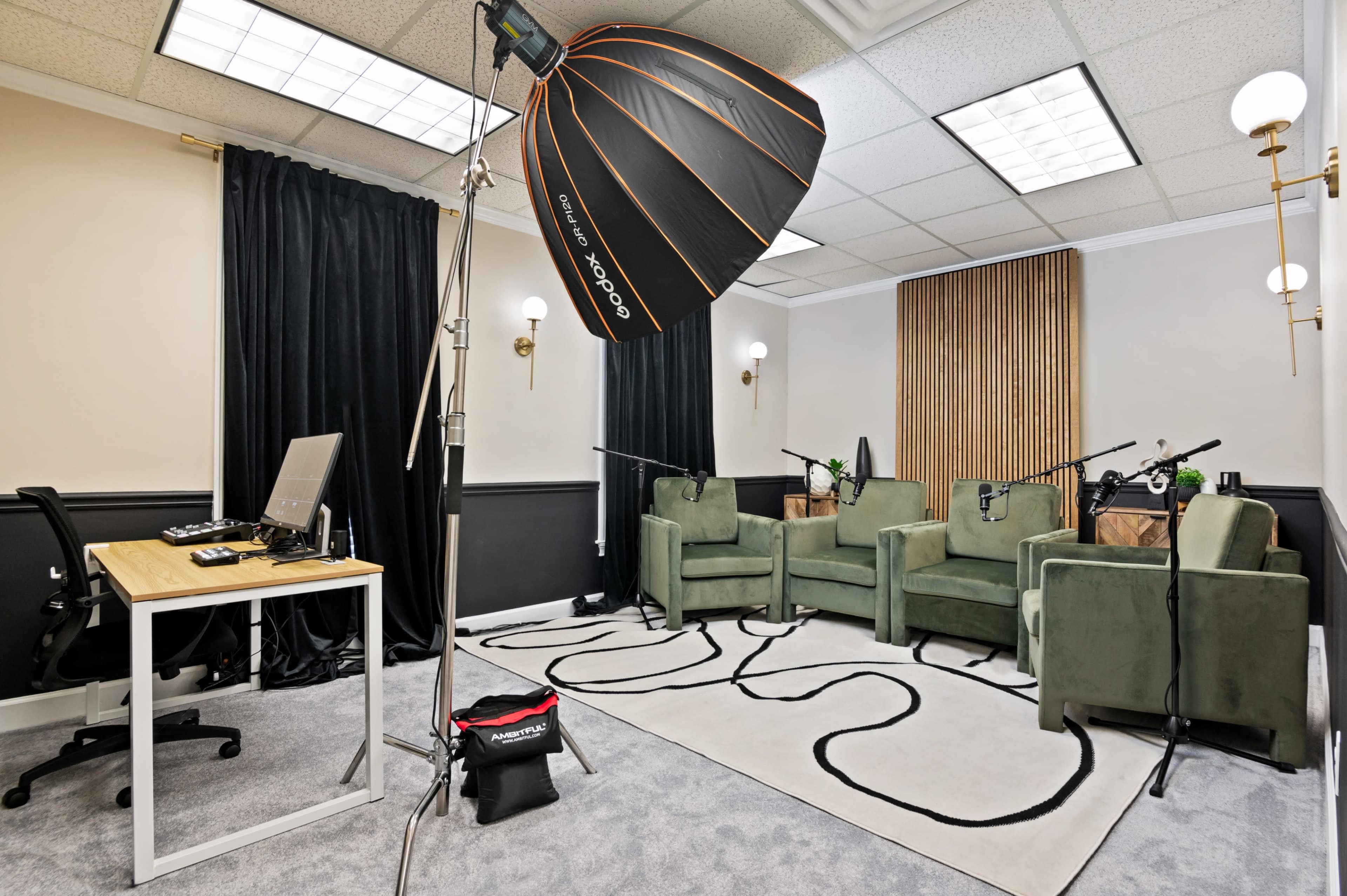 A recording studio is set up with three green chairs, professional microphones, and a desk in a well-lit room featuring black curtains and a patterned rug.