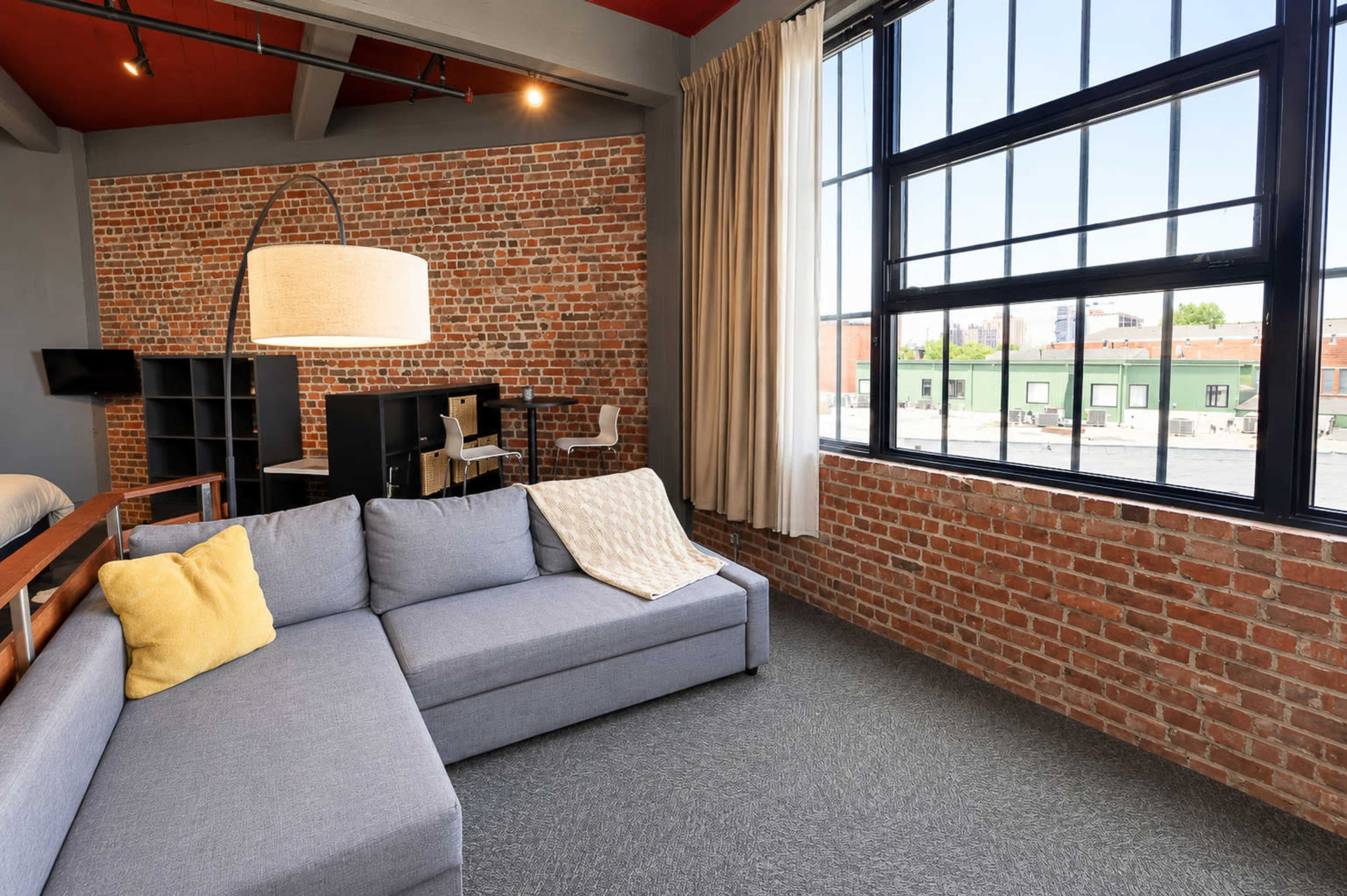 The image shows a modern living space with a gray sectional sofa, large windows, and exposed brick walls.