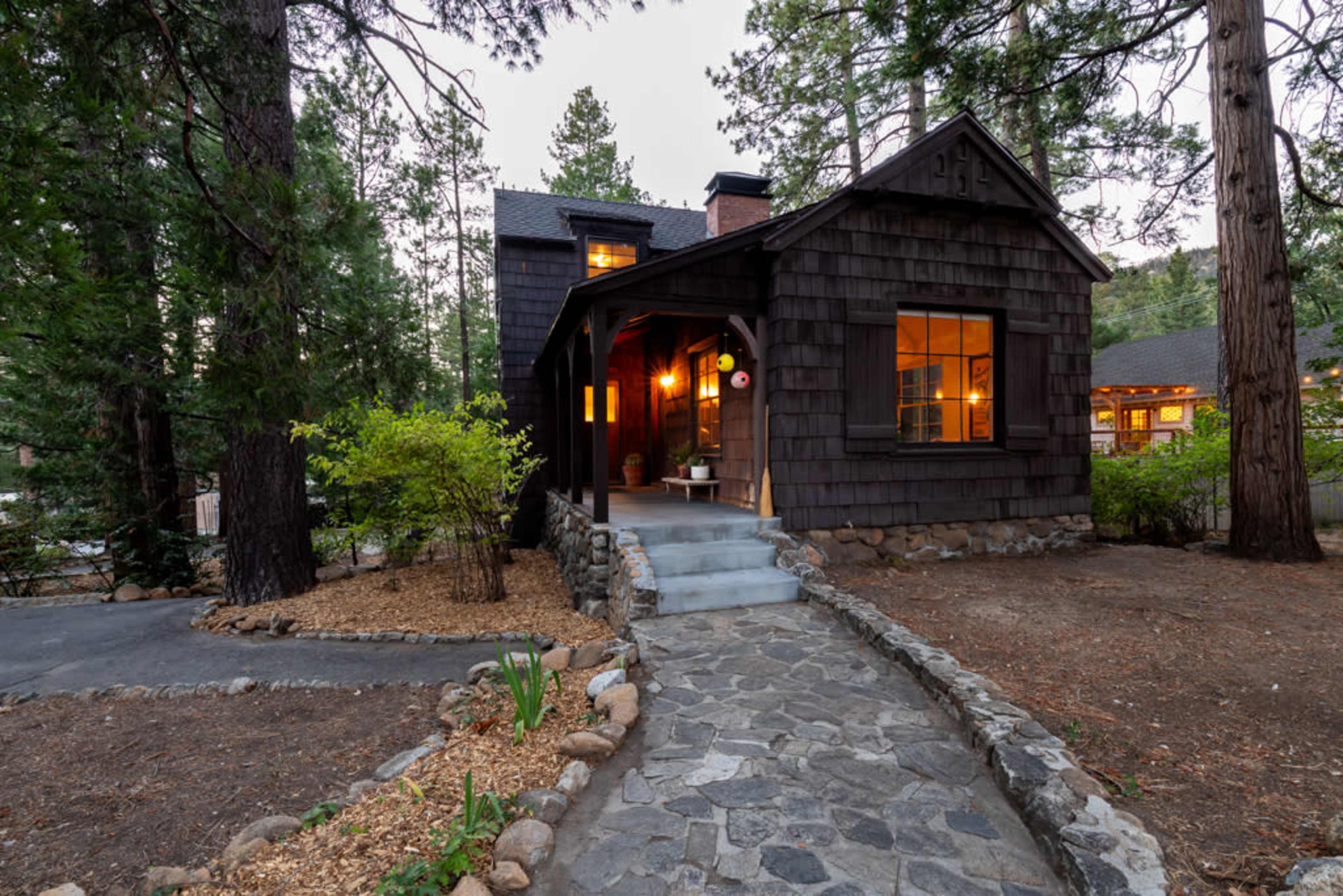 A wooden cabin with a stone pathway and illuminated windows is surrounded by tall trees in a forested area.