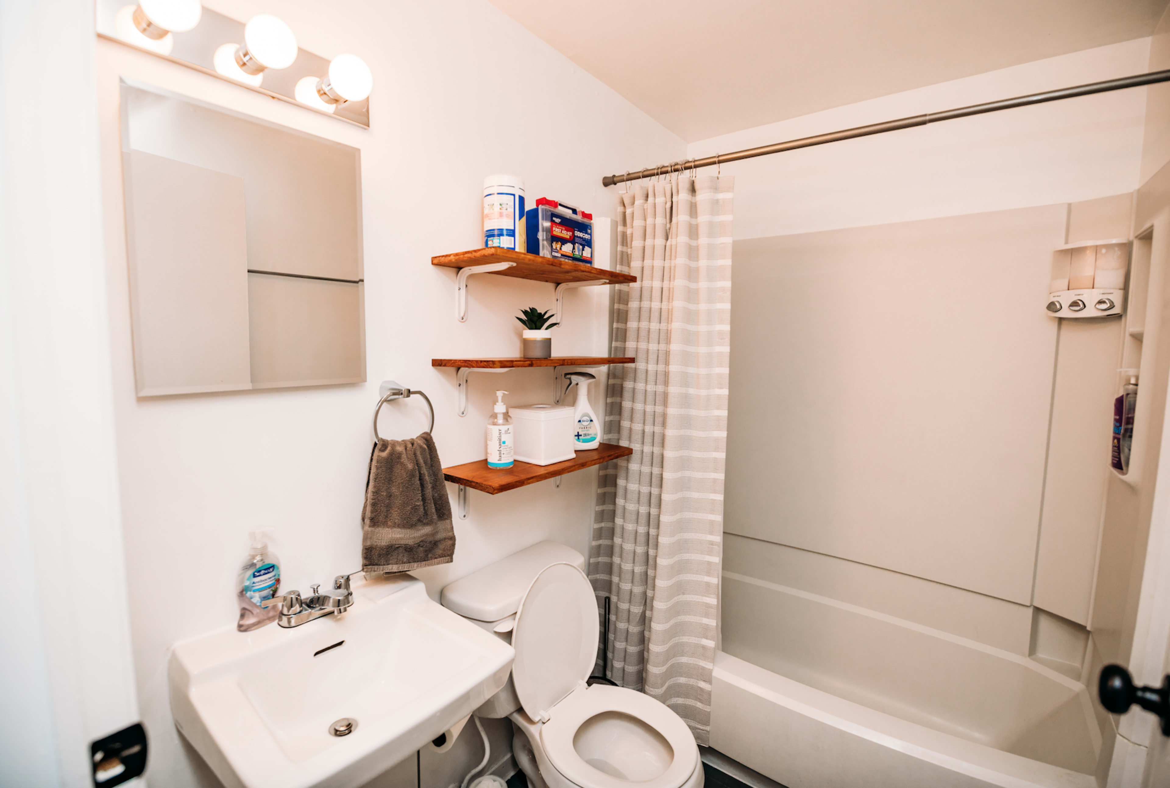 The image shows a bathroom featuring a sink, toilet, and a shower area with a towel rack and shelves holding toiletries.