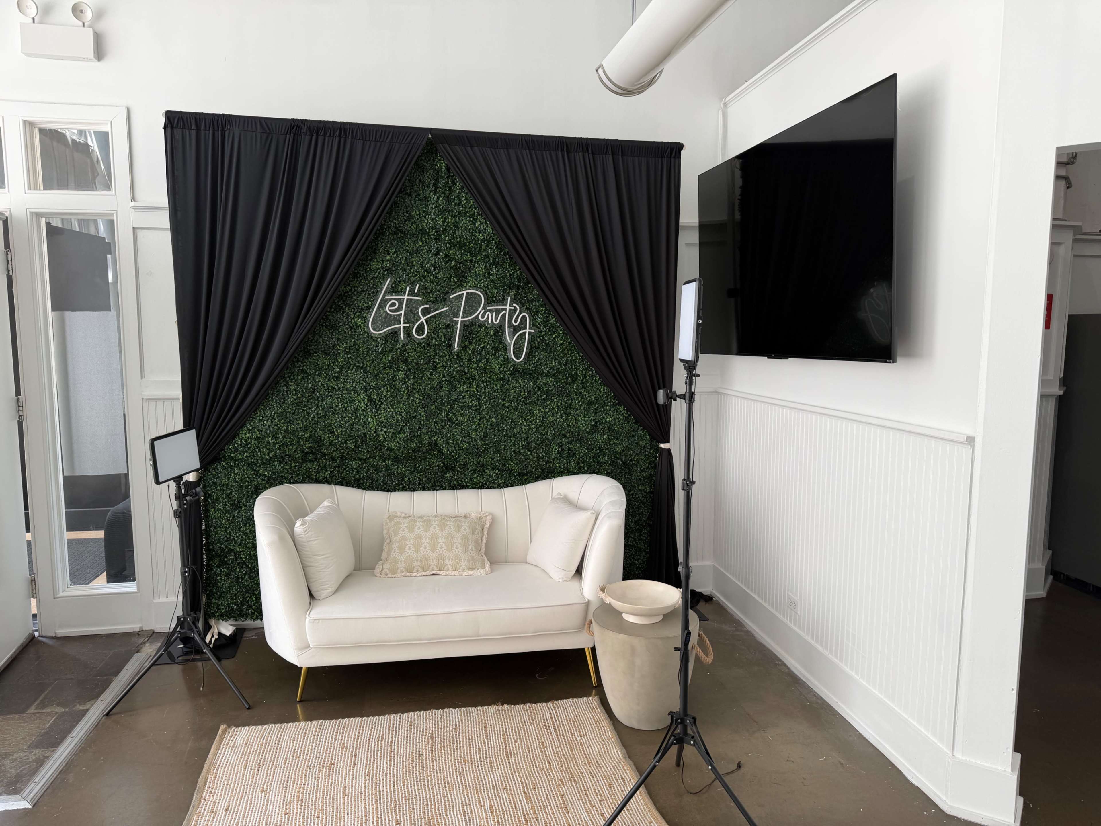 The image shows a white sofa in front of a green backdrop with the words "Let's Party" written in cursive, accompanied by two softbox lights and a television on the wall.