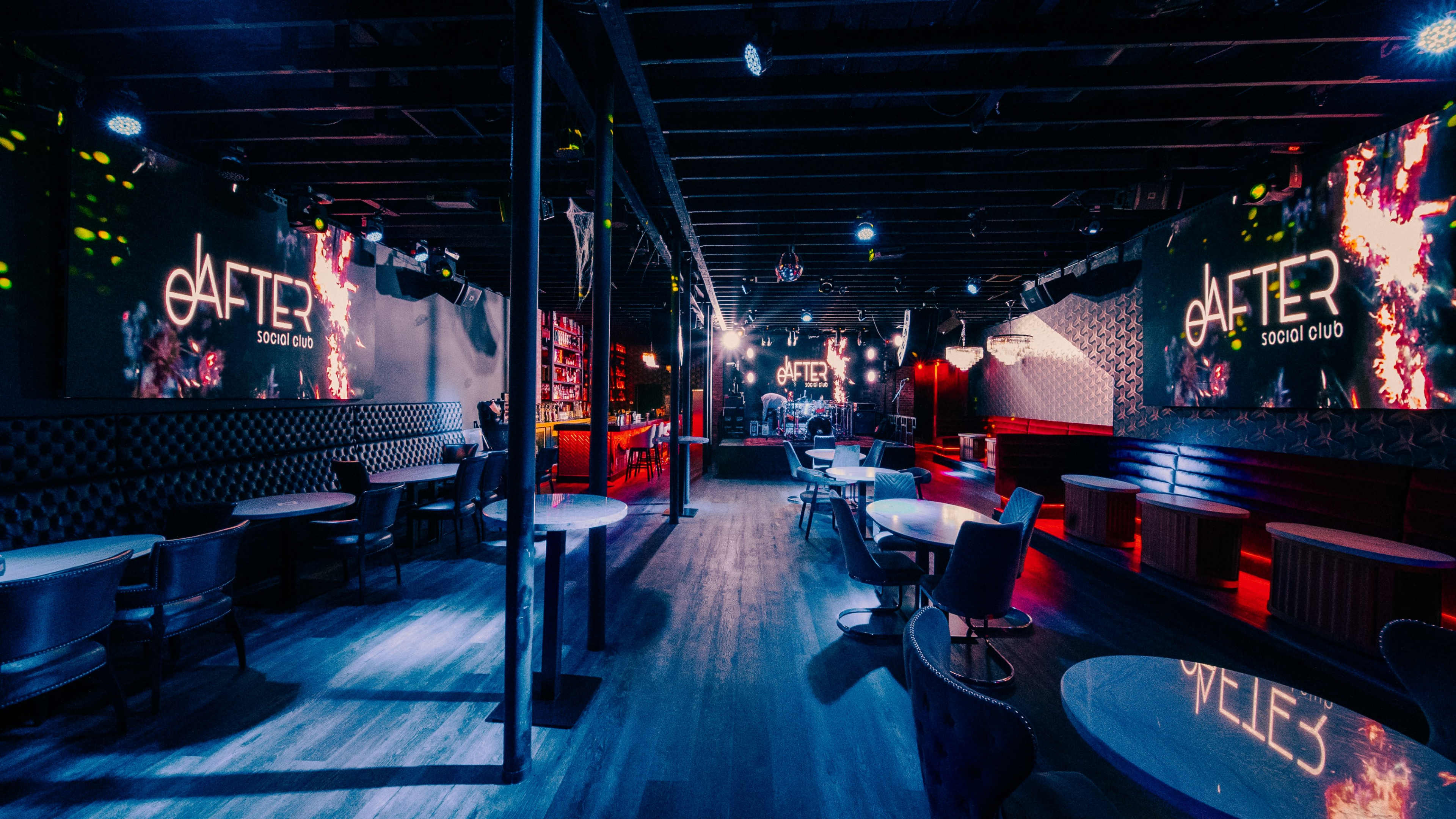 The image shows a dimly lit social club with modern decor, featuring round tables, a bar area, and large screens displaying the venue's logo.