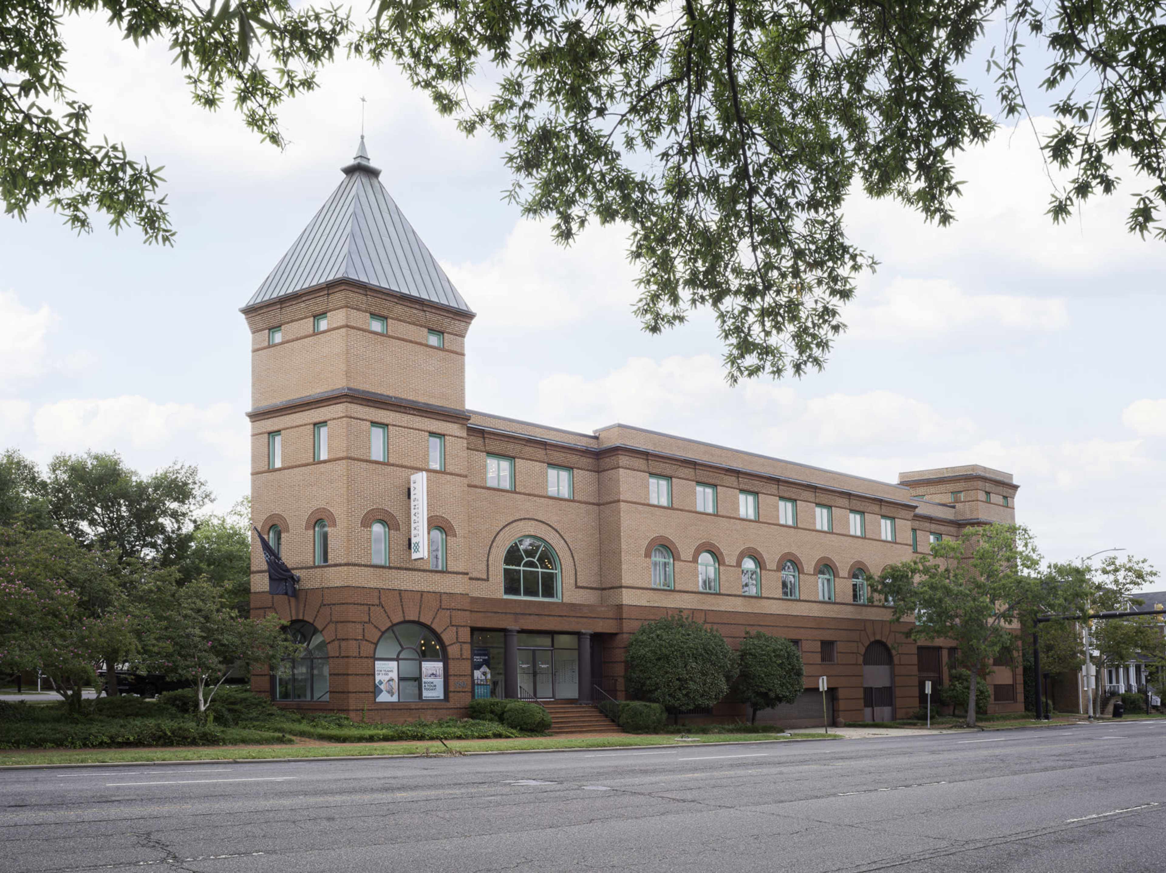 The image shows a three-story brick building with a triangular-shaped roof and large arched windows, situated on a tree-lined street.