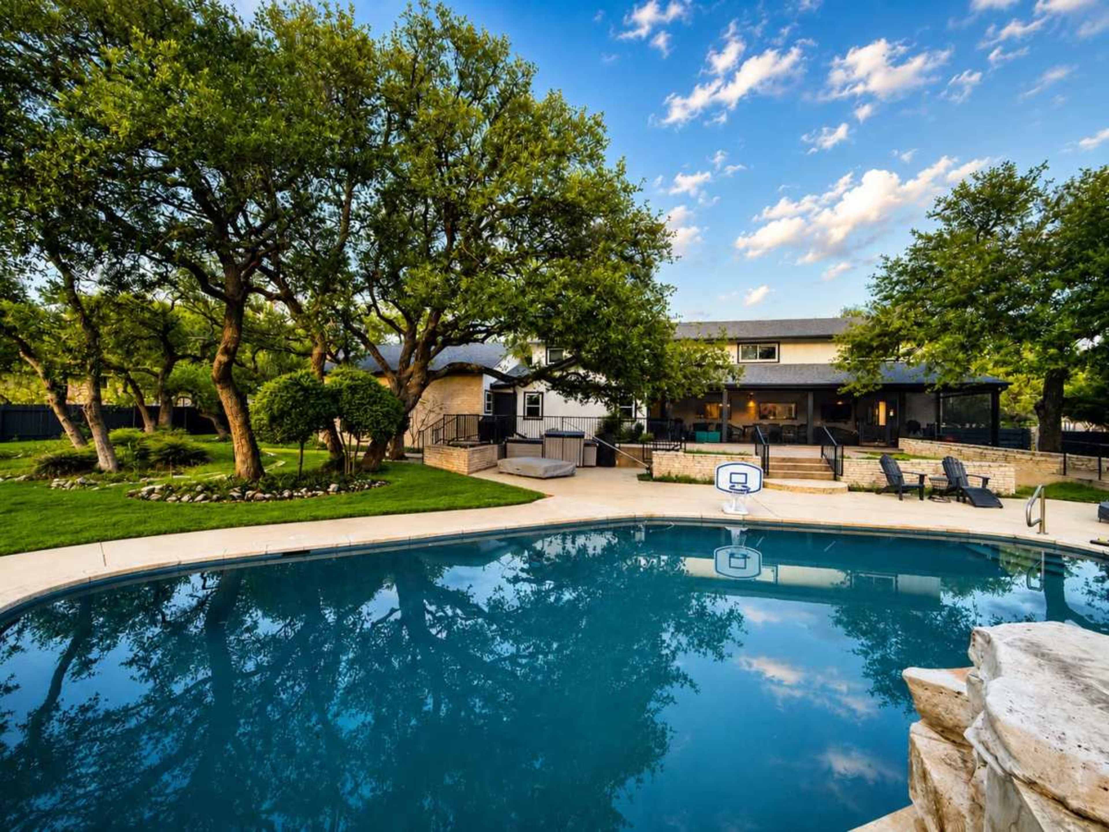 The image shows a backyard scene featuring a swimming pool surrounded by trees and a house in the background.