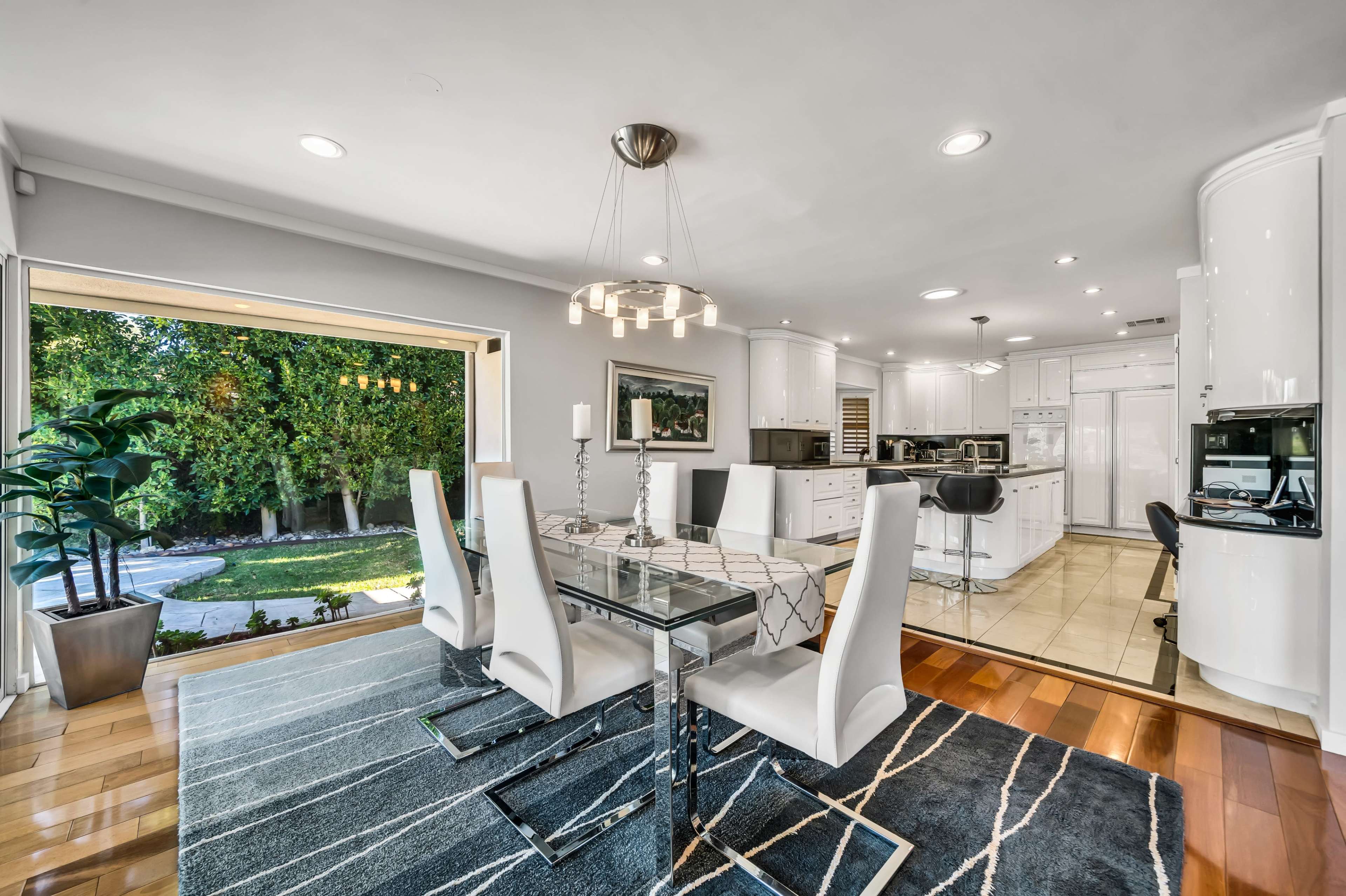 The image shows a modern kitchen and dining area featuring a glass dining table surrounded by white chairs, with a view of an outdoor space through large sliding doors.