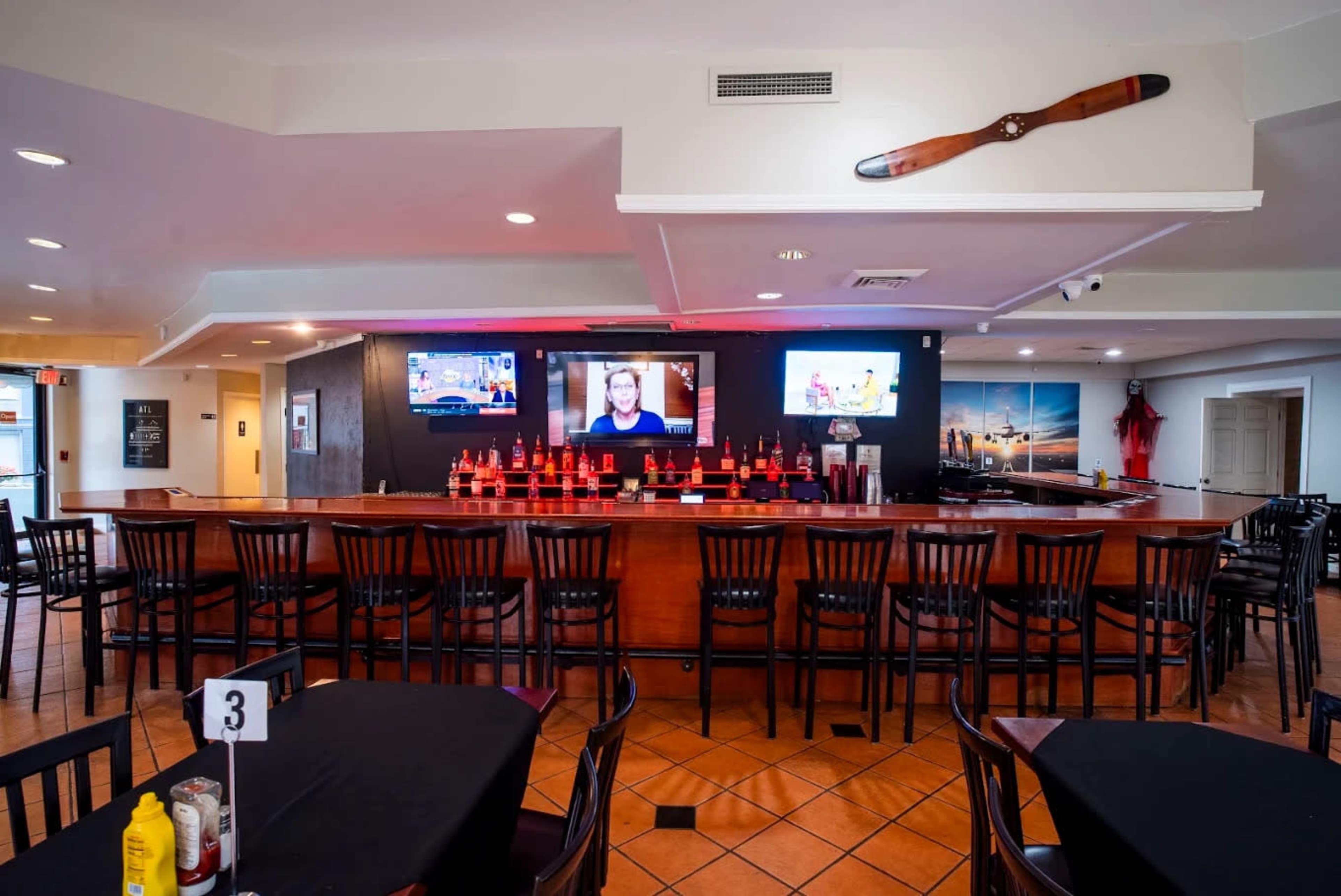 The image shows a bar area in a restaurant featuring a long counter with several chairs, two screens displaying content, and a decorative prop on the wall.