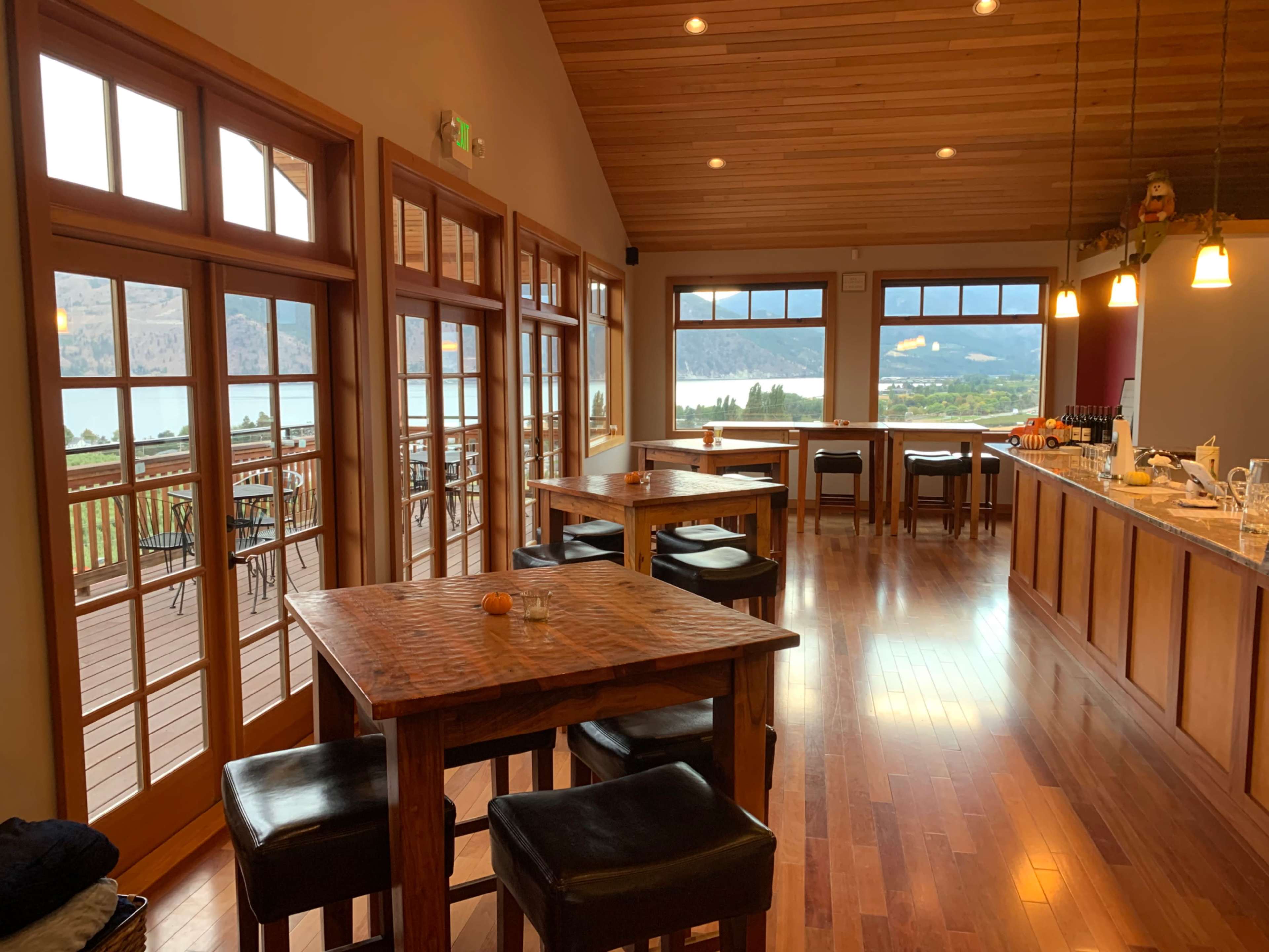 The image shows a spacious interior of a restaurant with wooden tables and chairs, large windows overlooking a scenic outdoor view, and a bar area in the background.